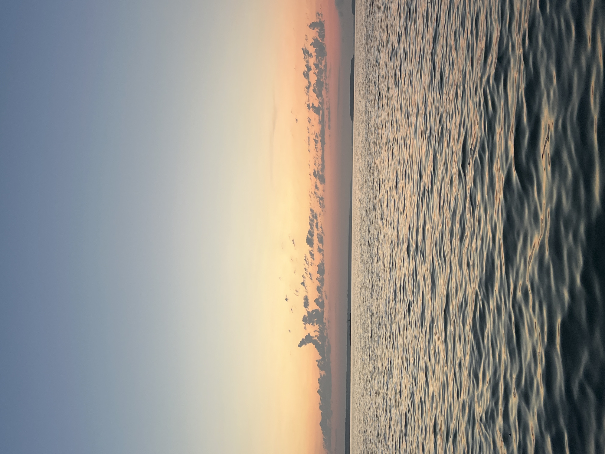 The image shows a serene body of water under a gradient sky at dusk. The sky transitions from a pale blue at the top to shades of orange and pink near the horizon, with scattered, wispy clouds. The water's surface is calm, with gentle ripples reflecting the colors of the sky. In the distance, a thin strip of land is visible, suggesting a coastline or the edge of a large lake. The overall scene evokes a peaceful and tranquil atmosphere. The location is specified as Novopichugovo, Russia.
