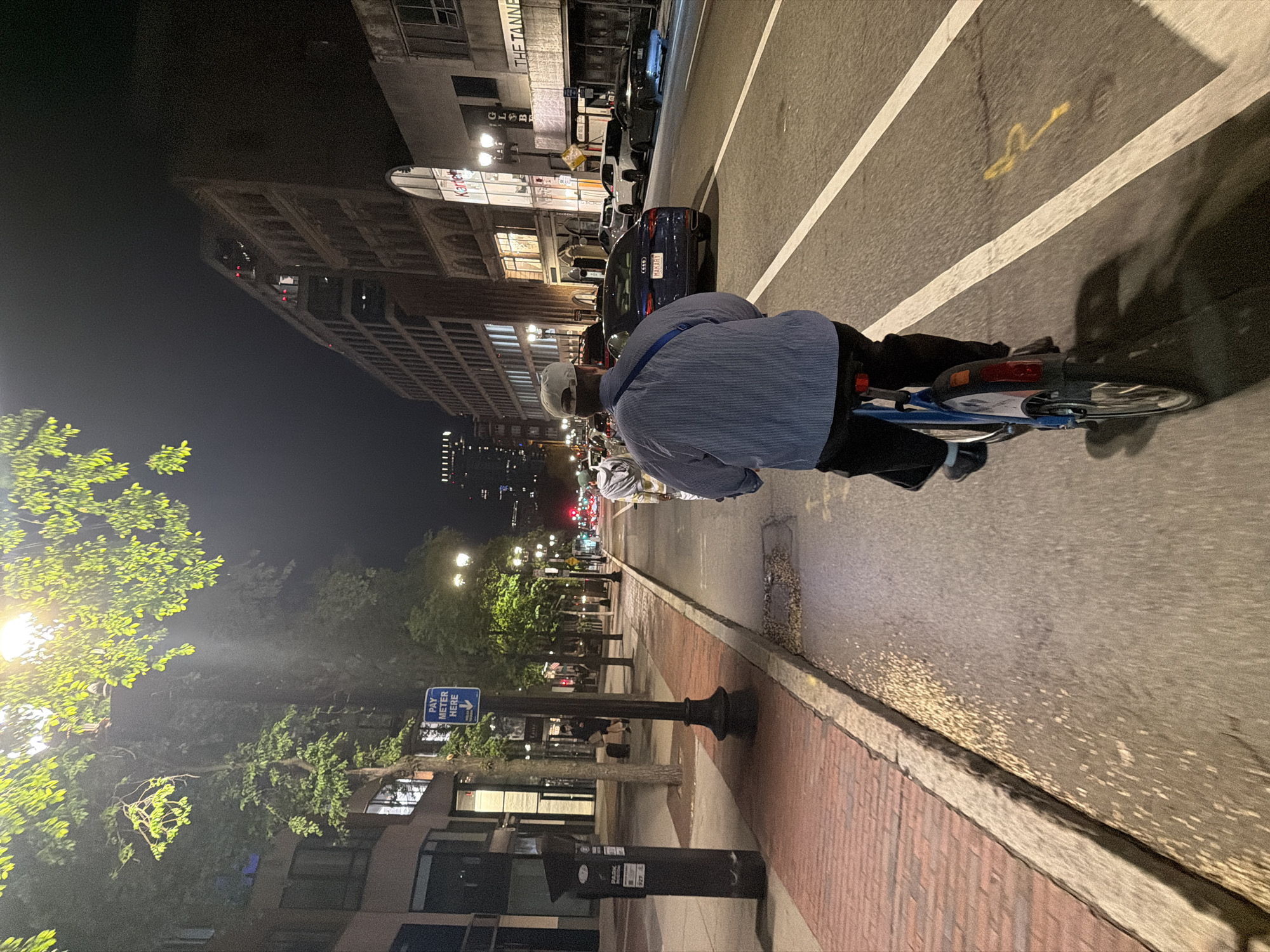 At night, the image depicts a man riding a blue bicycle on a city street in Boston, Massachusetts. He wears a light-colored baseball cap and a blue striped shirt. To his left is a brick sidewalk, a streetlamp, and a building bearing the sign "PAY METER HERE." Across the street are taller buildings and trees, all illuminated by streetlights. Several cars are visible in the adjacent lane. The street is divided by painted white lines.