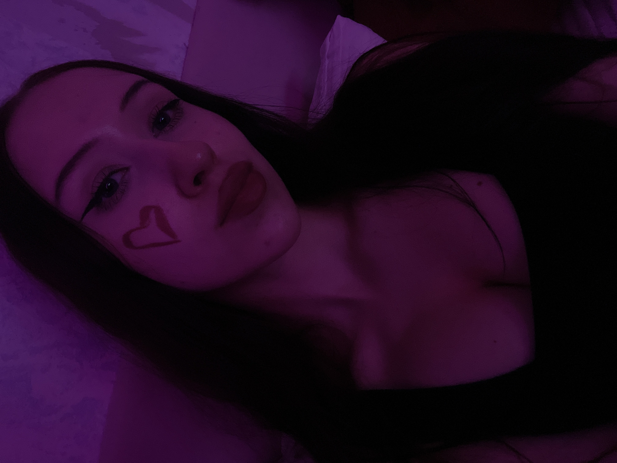 The image is a close-up selfie of a young woman with long dark hair. She is wearing a dark top and has a heart shape drawn on her left cheek. Her makeup includes winged eyeliner and red lipstick. The lighting in the image is predominantly purple, suggesting an artificial light source. The background appears to be a wall or fabric, with some texture visible. There is no indication of the city of Votkinsk, Russia, or any specific location within it. The image focuses solely on the woman's face and upper body.