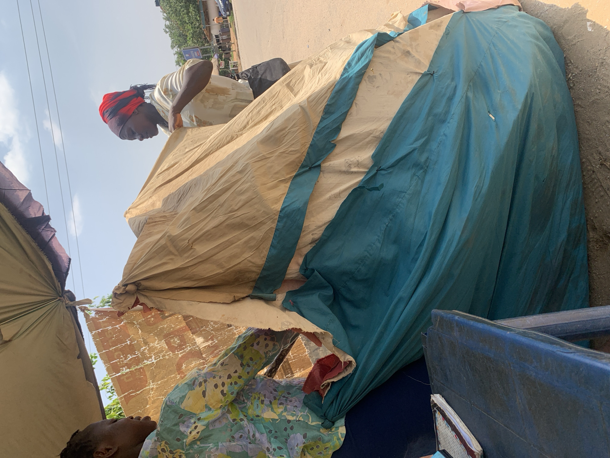 Two women are in a market stall in Abuja, Nigeria, engaged in setting up or packing away a large, layered fabric structure. The structure appears to be made of several tarps or sheets of material in beige and teal, with some red fabric showing. The woman on the left, wearing a green and blue floral patterned shirt, is standing and looking at the fabric. The woman on the right, wearing a light-colored shirt and a red and black headscarf, is also standing and interacting with the fabric. Behind them, a sandy ground is visible, and a blue plastic table with a notebook on it is in the foreground. In the background, there are trees, buildings, and a street with vehicles and people, suggesting an outdoor urban environment. The sky is visible and appears partly cloudy. The scene captures a moment of work within a bustling marketplace.