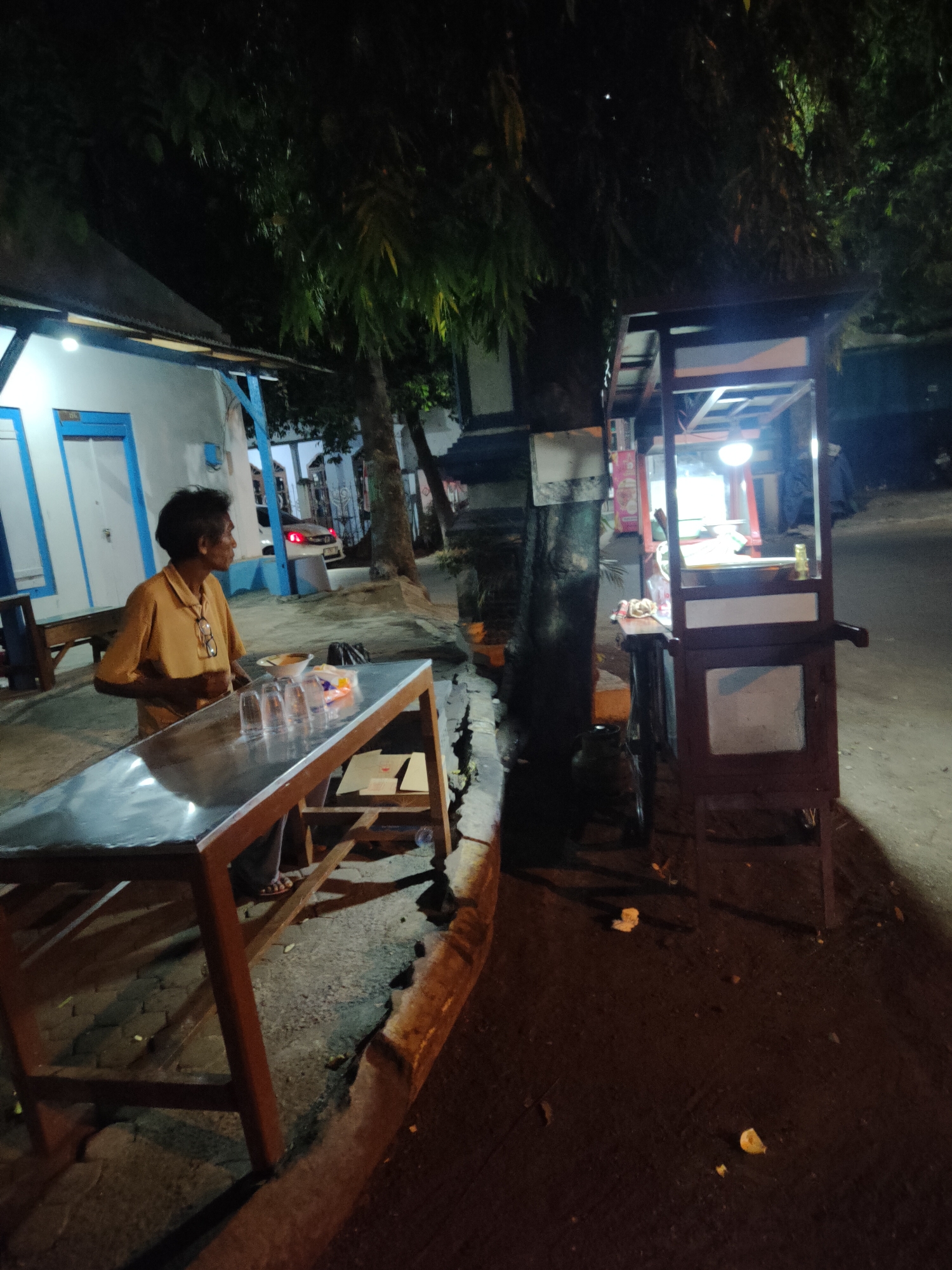 A man in a yellow shirt stands beside a stainless steel table with several glasses and a bowl on it. He is looking to his right. In the background, a food cart with a light illuminates its interior. The scene takes place at night on a street in Bangil, Indonesia, with trees and buildings visible.