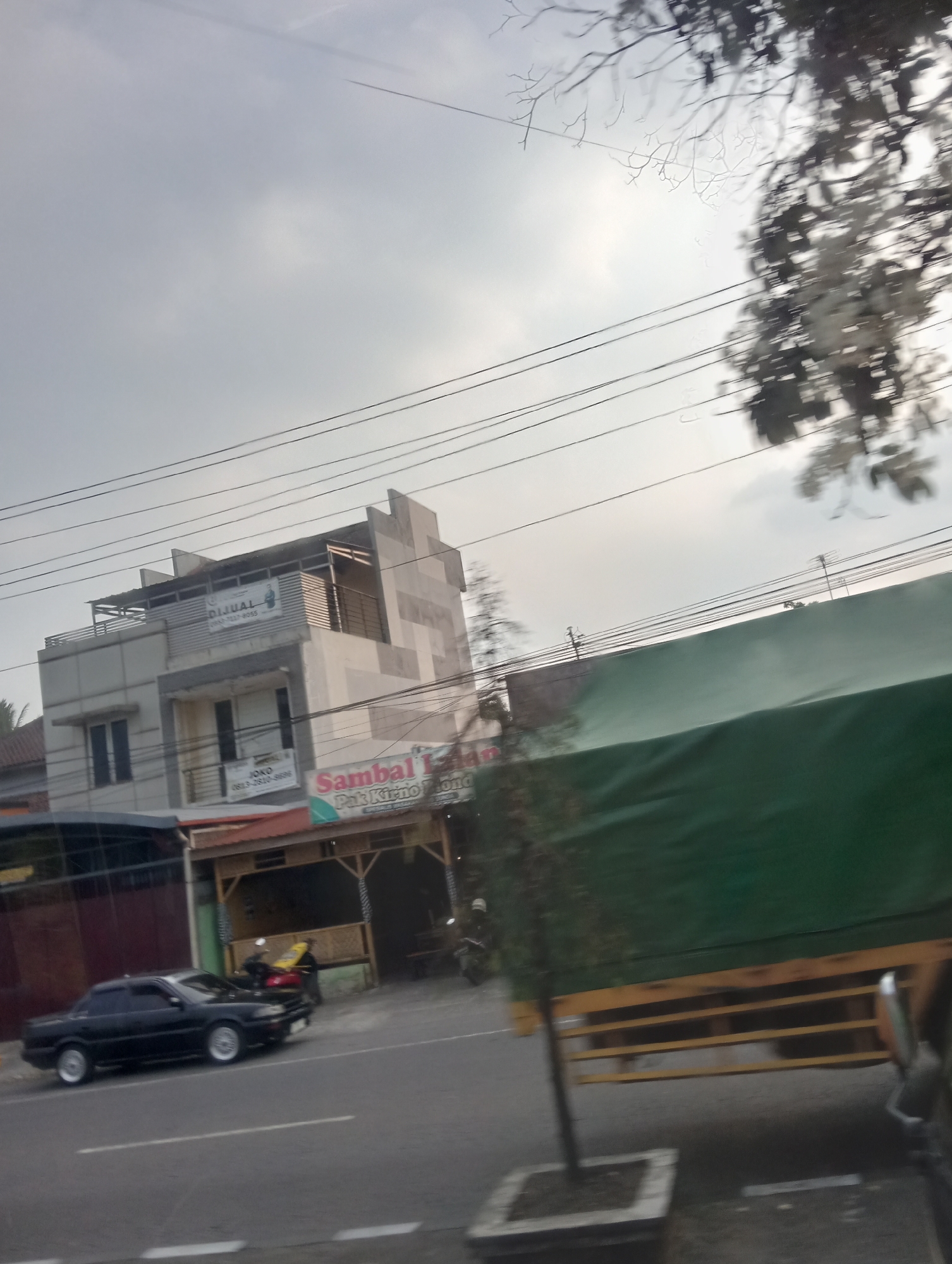 A street-level, outdoor, medium-shot captures a cloudy day in the city of Danurejo, Indonesia, with a moving vehicle perspective. In the mid-ground, a multi-story building with a white and grey facade stands, featuring a sign that reads "DIJUAL" (For Sale) and the phone number "0813-7817-8055." Below this, another sign indicates "JOYO" and a phone number. To the right of this building, a smaller structure with a bamboo-like facade and a red roof is visible, with a sign that says "Sambal Pak Kirno." In the foreground, a black sedan is parked on the right side of the road, and a large truck with a green tarp covers its cargo is partially visible. The environment includes power lines crisscrossing the sky, some blurred trees, and the asphalt road.