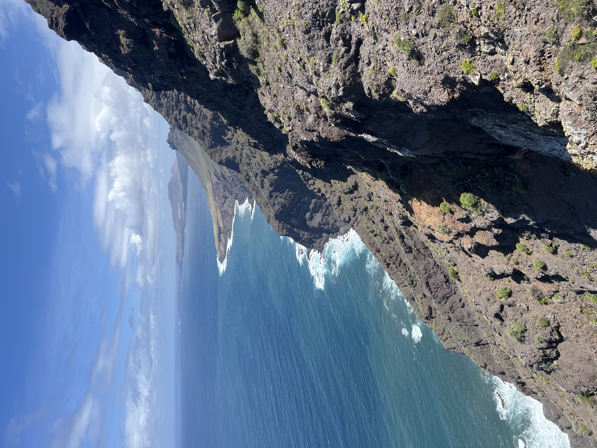 The image shows a high-angle view of the rugged coastline of Tirma, Spain. Steep cliffs, covered in sparse vegetation, drop dramatically into the deep blue ocean. White waves crash against the base of the cliffs, creating sea foam. The sky is partly cloudy with blue patches. In the distance, other land formations are visible along the coast. There are no people visible in the image.