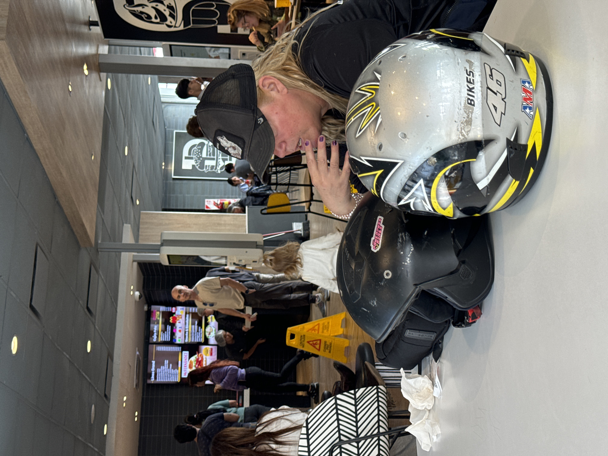 This image captures a candid moment inside a modern fast-food restaurant, likely in Buenos Aires, Argentina, given the Spanish text on the menu boards. The setting features sleek gray and wooden wall panels, illuminated by recessed lighting.
In the immediate foreground, a woman with long blonde hair, wearing a black cap with a wolf emblem and a black shirt, leans over a white table. Her hand, adorned with purple nail polish and a silver bracelet, rests near her face. Two motorcycle helmets are prominently displayed on the table beside her. One helmet is silver/gray with black and yellow accents, featuring the text "BIKES," "46," and an "AMA" logo. The other is a black helmet with a partially visible pink sticker that appears to say "CALIFORNIA" or similar. White crumpled tissues are also on the table.
In the midground, several individuals are engaged in ordering or waiting. A bald man in a light-colored shirt stands near the order counter, where digital menu boards display various food items in Spanish, such as "Lomo," "Grand," and "McFRIES." Further along the queue, a woman with long dark hair and a purple top looks at the menu. On the floor, a bright yellow "CAUTION WET FLOOR" sign is visible.
The background shows more patrons seated at tables, and a large, stylized graphic of an animal head is displayed on an overhead panel. The overall activity suggests a busy, casual dining environment where people are getting food and relaxing. The presence of motorcycle helmets implies the foreground subjects may have arrived by motorcycle.