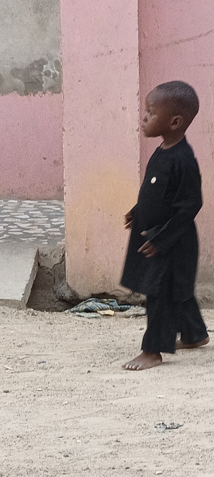 An outdoor scene in Rangaza, Nigeria, features a young, dark-skinned child captured in a profile view, facing towards the left side of the frame. The child is dressed in a traditional-style black long-sleeved tunic and matching trousers, with a small, circular white badge pinned to the left side of their chest. They are barefoot, walking or standing on a sandy, dusty ground speckled with small pebbles and debris.
The background consists of a textured wall, primarily painted pink, with patches where the paint appears faded or chipped, revealing lighter, possibly gray, sections underneath. To the child's left, a rough, gray concrete structure, possibly a step or foundation, is visible at the base of the wall. Near this structure and on the ground lies a crumpled piece of blue and white striped fabric.
The lighting indicates it is daytime. The child's expression is calm and focused, gazing ahead. No legible text is visible on the badge or elsewhere in the image.