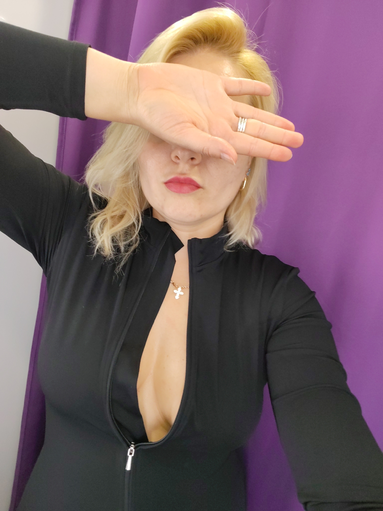 A woman with blonde hair, wearing a black zippered dress and silver rings, covers her face with her hand. She is wearing gold earrings and a cross necklace. The background is a vibrant purple curtain. The image appears to be taken indoors, possibly in a changing room or a studio, given the purple curtain and the plain backdrop. There is no visible text in the image. The lighting suggests it could be daytime, or artificial lighting is used indoors. The woman's lips are painted pink, and her expression, obscured by her hand, cannot be determined.
