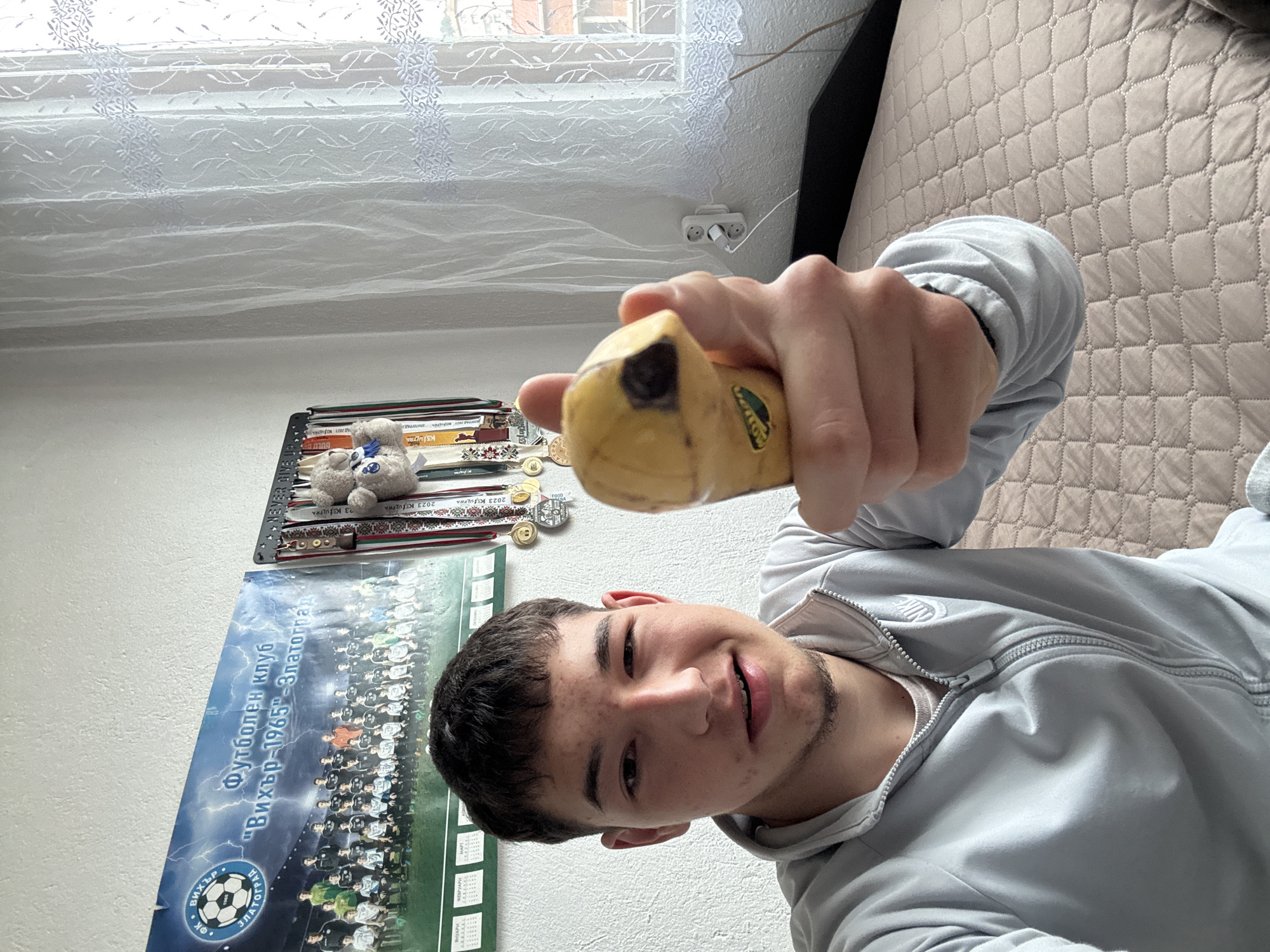 The image shows a young man holding a partially eaten banana in his right hand, presumably taking a selfie. The background includes a wall with a poster of a football club named "Вихър-1965"-Златоград (Vikhar-1965-Zlatograd), likely from the city of Zlatograd, Bulgaria. Above the poster, there is a shelf with a teddy bear and what appears to be medals or coins. A window with a sheer curtain is visible in the upper part of the image, along with a power outlet. The man is wearing a gray Nike tracksuit jacket. A bed with a quilted pattern is also present.