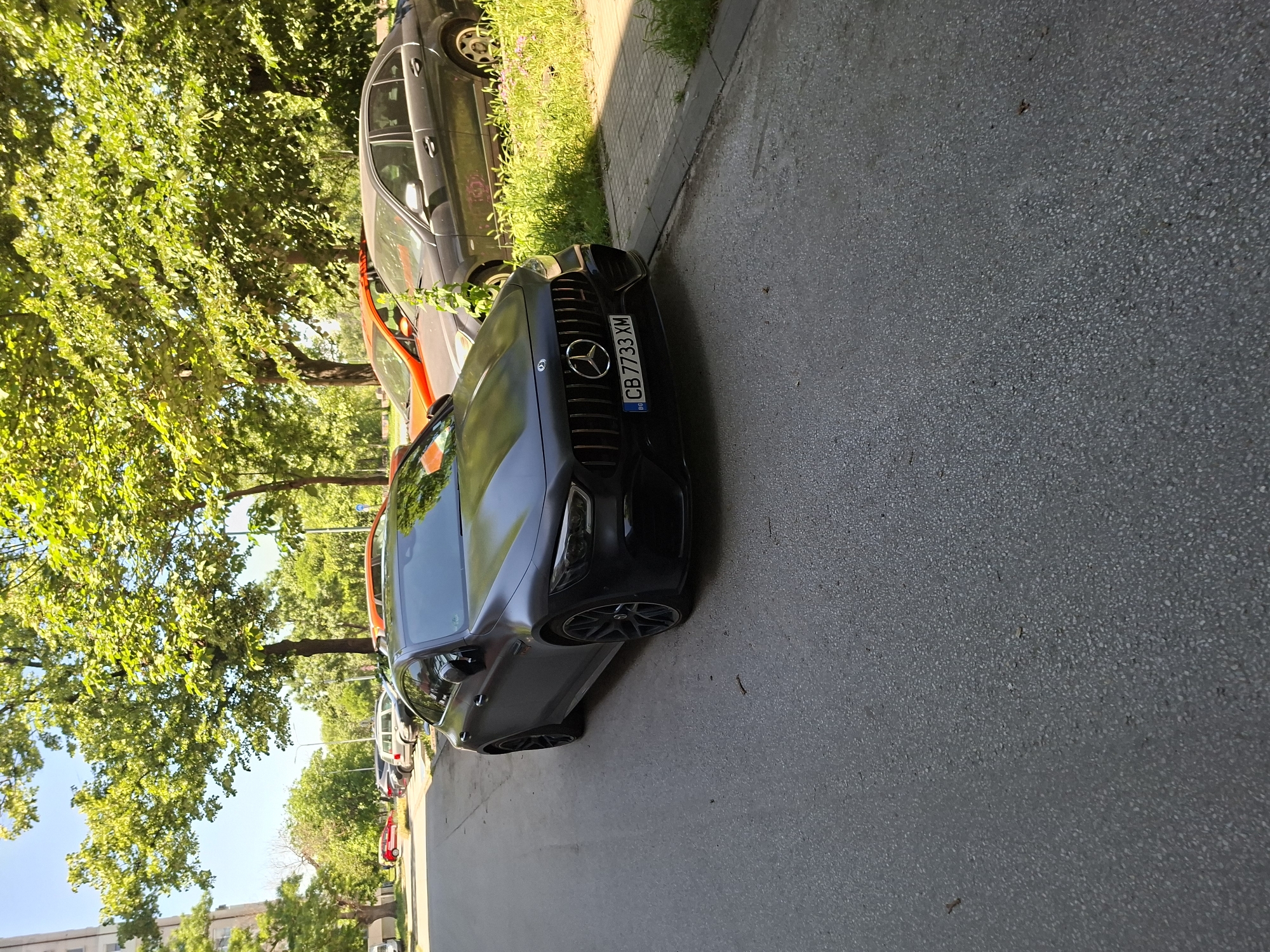 The image shows a black Mercedes-Benz car parked on the side of a street. The car has a license plate that reads "CB 7733 XM," indicating it is registered in Bulgaria, specifically in Burgas, where the "CB" prefix is used. The street is lined with trees on the left side, and there are other cars parked in the distance. The location appears to be a residential area in Plovdiv, Bulgaria, given the types of cars parked along the street and the general layout. The scene is during daylight hours with bright sunlight.