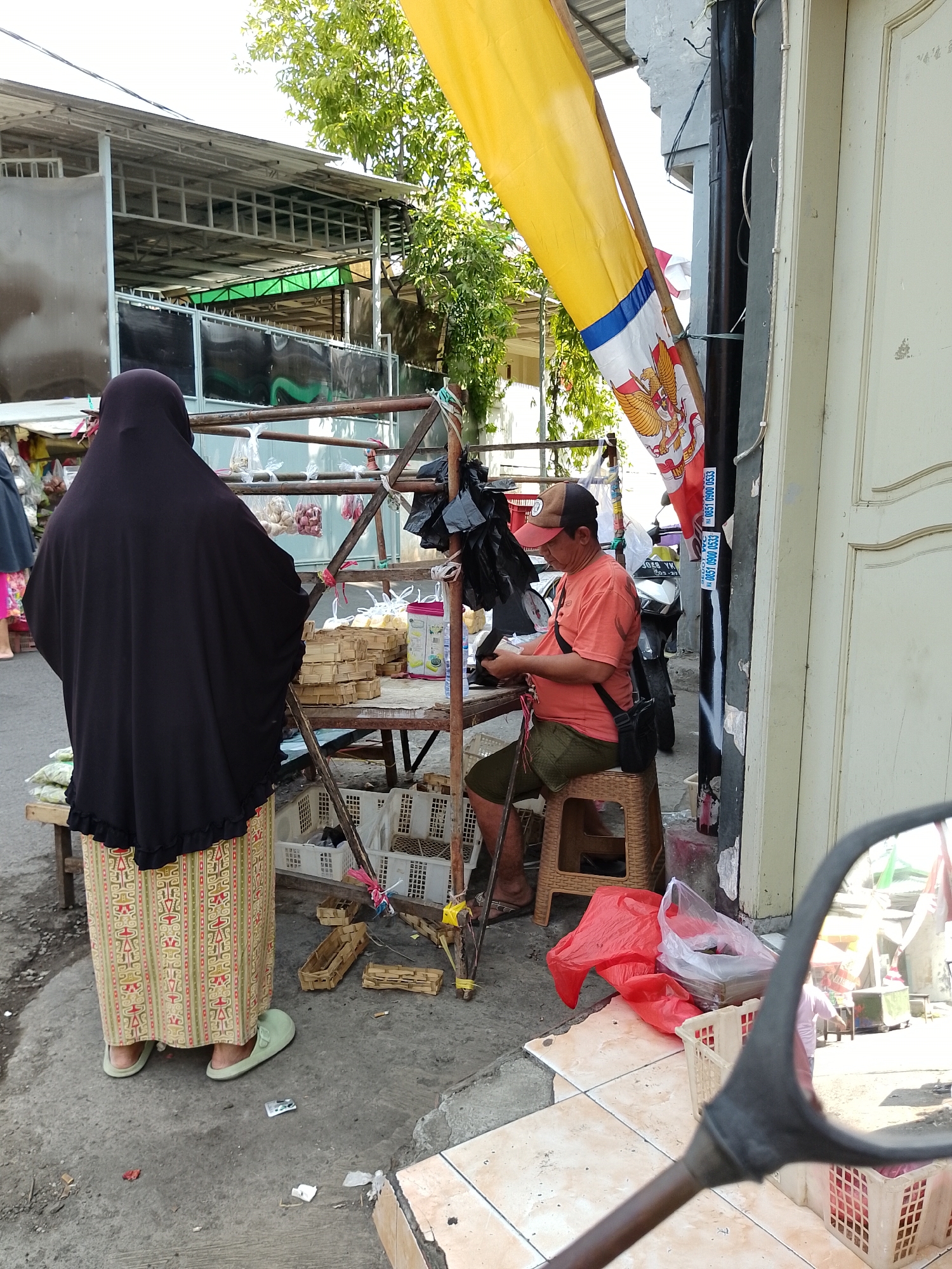 In Surabaya, Indonesia, on a sunny day, a scene unfolds at a roadside market. A woman wearing a black hijab and a patterned skirt stands with her back to the camera, facing a market stall. She is wearing light green slip-on shoes. To her right, a man in an orange t-shirt and green shorts sits on a woven stool, looking down and appearing to be working with something in his hands. He has a black bag slung across his chest and is wearing a baseball cap. The market stall itself is constructed of metal and wood, with various goods displayed. Baskets and plastic bags containing produce like onions and what appear to be crackers are visible. In the background, a yellow flag with blue and red stripes and an emblem is attached to a pole. A motorcycle is parked nearby. The ground is paved with concrete and tiles. Trash, including a red plastic bag, is scattered on the ground. The overall impression is one of a bustling, everyday market scene in an Indonesian city.