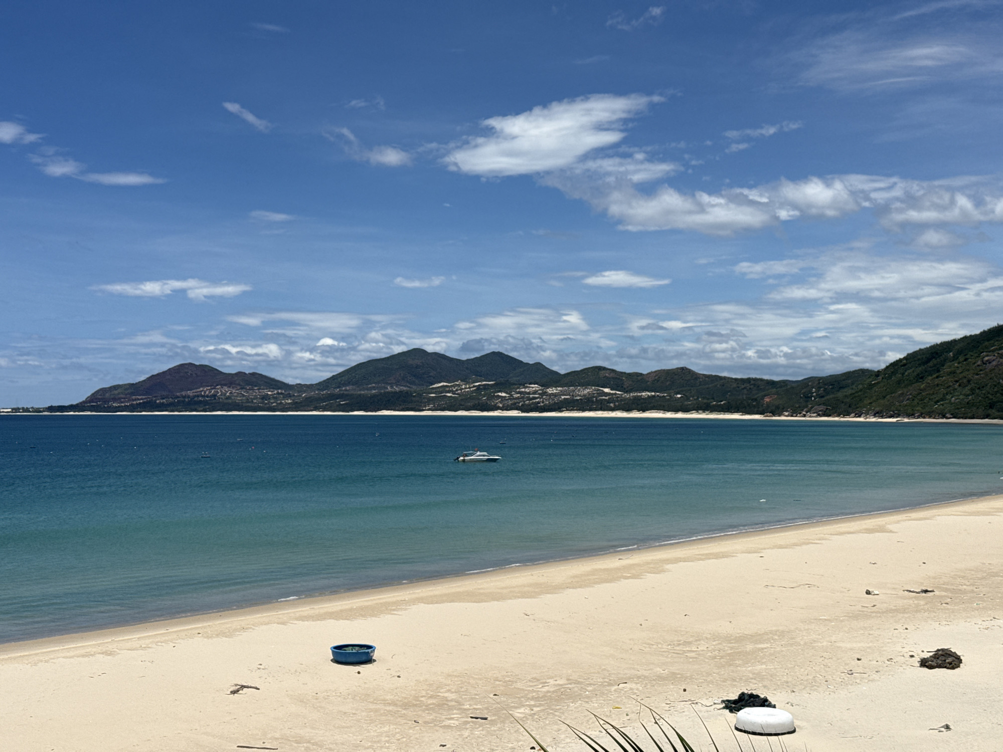 A wide shot captures a tranquil beach scene under a partly cloudy blue sky. The sand is a light beige, with a few scattered pieces of debris, including a blue bucket and some white and dark organic material. The ocean stretches out in front of the beach, transitioning from a pale turquoise near the shore to a deep teal further out. A small white motorboat is visible on the water, about a third of the way from the left of the frame. In the distance, a range of green, tree-covered mountains forms the horizon, with a town nestled along their foothills. The overall impression is one of a peaceful, natural coastal landscape.