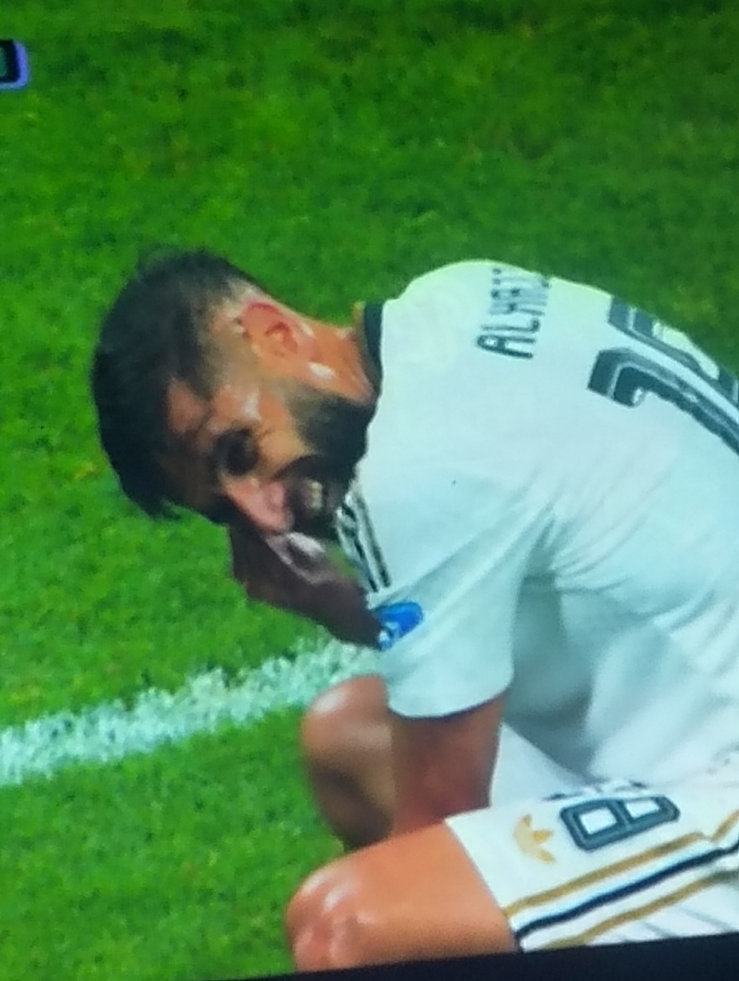 A man, likely a soccer player, is crouched on a bright green field, his head bowed and his hand touching his face. He is wearing a white jersey with black lettering that appears to read "ALHAZ" on the back and a number "10" on his right sleeve. The jersey also has gold and black stripes on the sleeves and shorts, and an Adidas logo on his left arm. The field has a white line marking its boundary. The man's skin is flushed, suggesting exertion or distress. There is no visible indication of the city of Payagaboh, Indonesia.