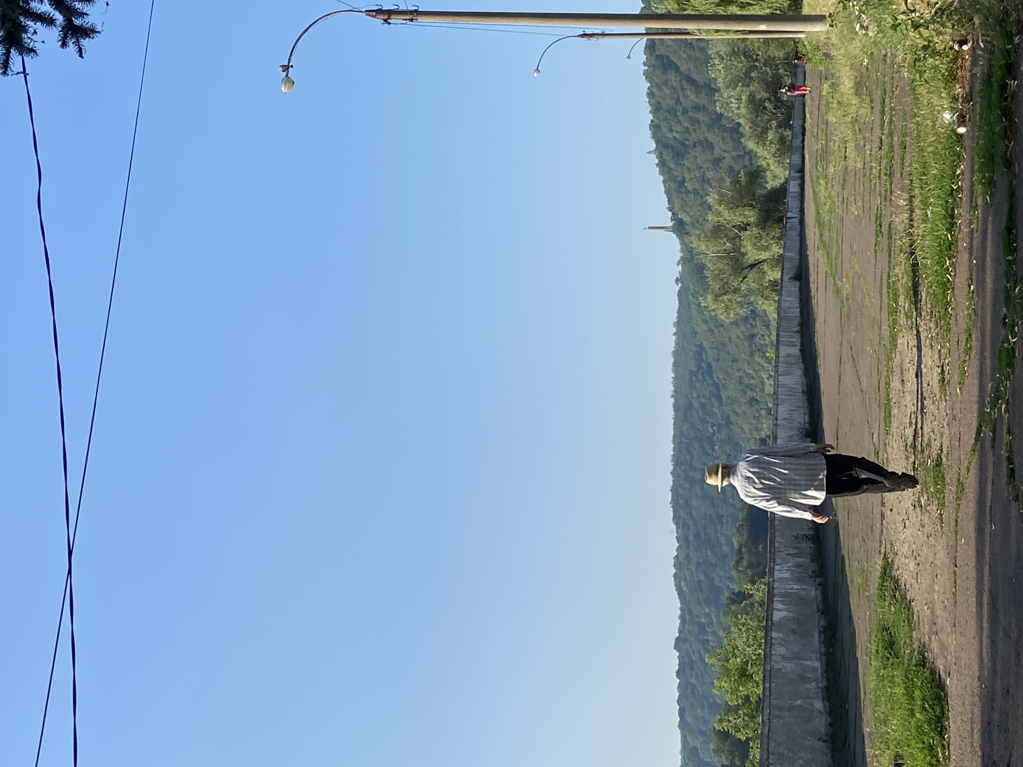 An older man wearing a straw hat, a striped collared shirt, and dark trousers walks away from the viewer on a paved path. He is walking towards a low concrete wall, behind which is a lush green hillside covered in trees. The sky is a clear, bright blue. To the right of the path are lampposts, and in the distance, a tall, thin structure resembling a monument or tower is visible on top of the hill. A person in red clothing is seen walking on the path further in the distance. The scene appears to be an outdoor recreational area or park.