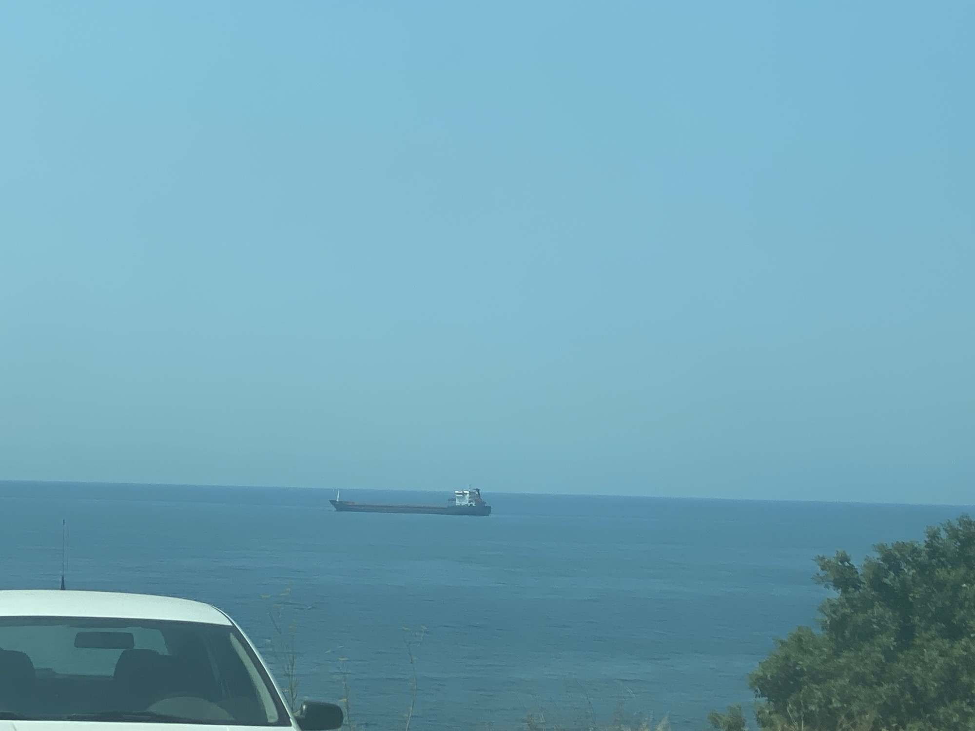 The image captures a wide view of the Adriatic Sea with a large cargo ship in the distance. The sky is a clear, light blue. In the foreground, the roof and windshield of a white car are visible, suggesting the photo was taken from inside or beside a vehicle. To the right, lush green foliage borders the scene. The vantage point indicates a location overlooking the sea, consistent with areas along the coast of Bar, Montenegro.