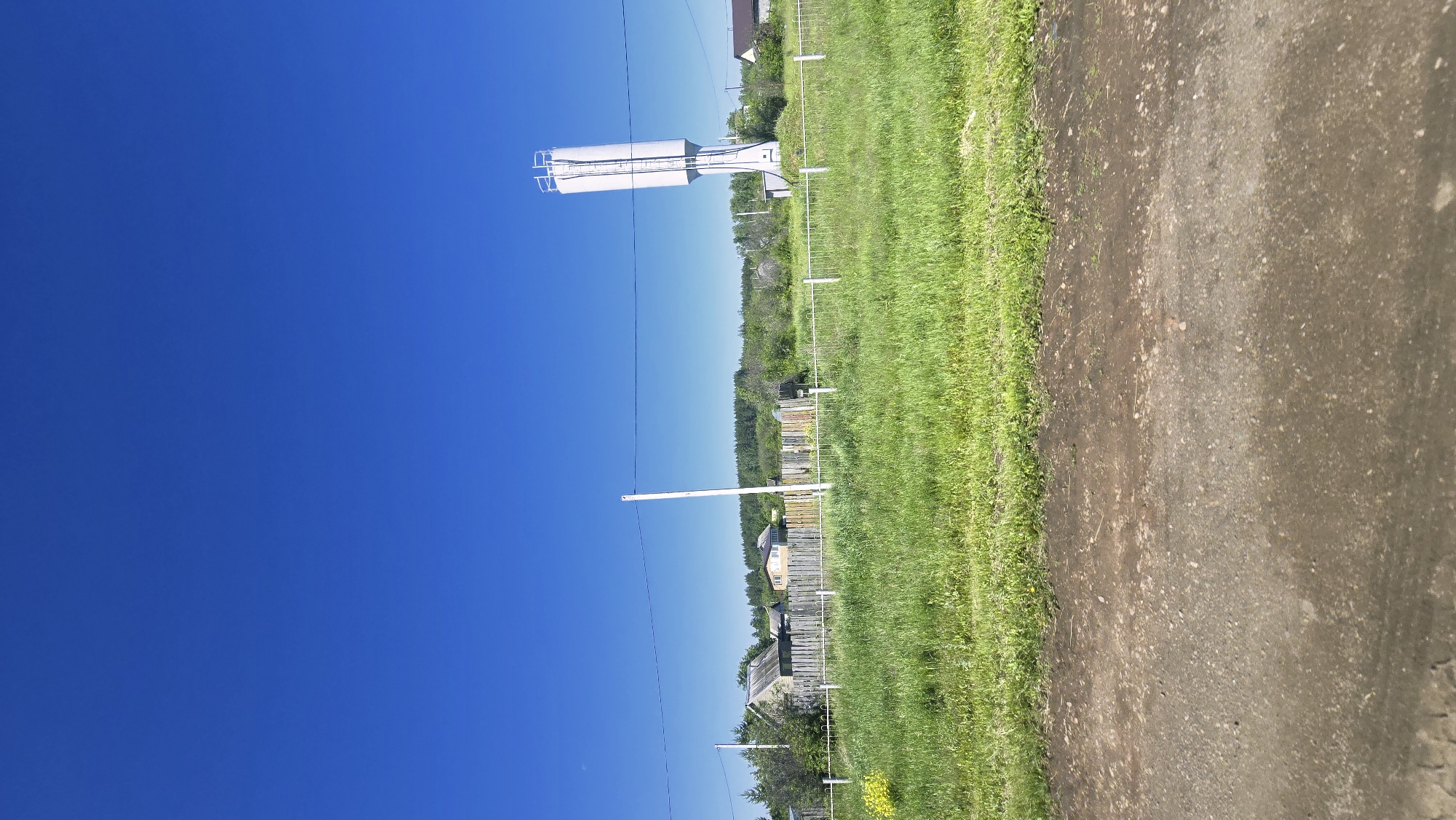 The image captures a rural scene under a clear blue sky. In the foreground, a dirt road runs horizontally across the bottom of the frame. Beyond the road, a strip of lush green grass extends towards a weathered wooden fence. Behind the fence, several houses with wooden fences and roofs are visible amidst trees and vegetation. To the far right of the scene, a tall, cylindrical white water tower stands prominently against the sky. Power lines are strung across the middle ground. The overall impression is that of a quiet, rural settlement. The location is identified as Актюбинский, Russia.