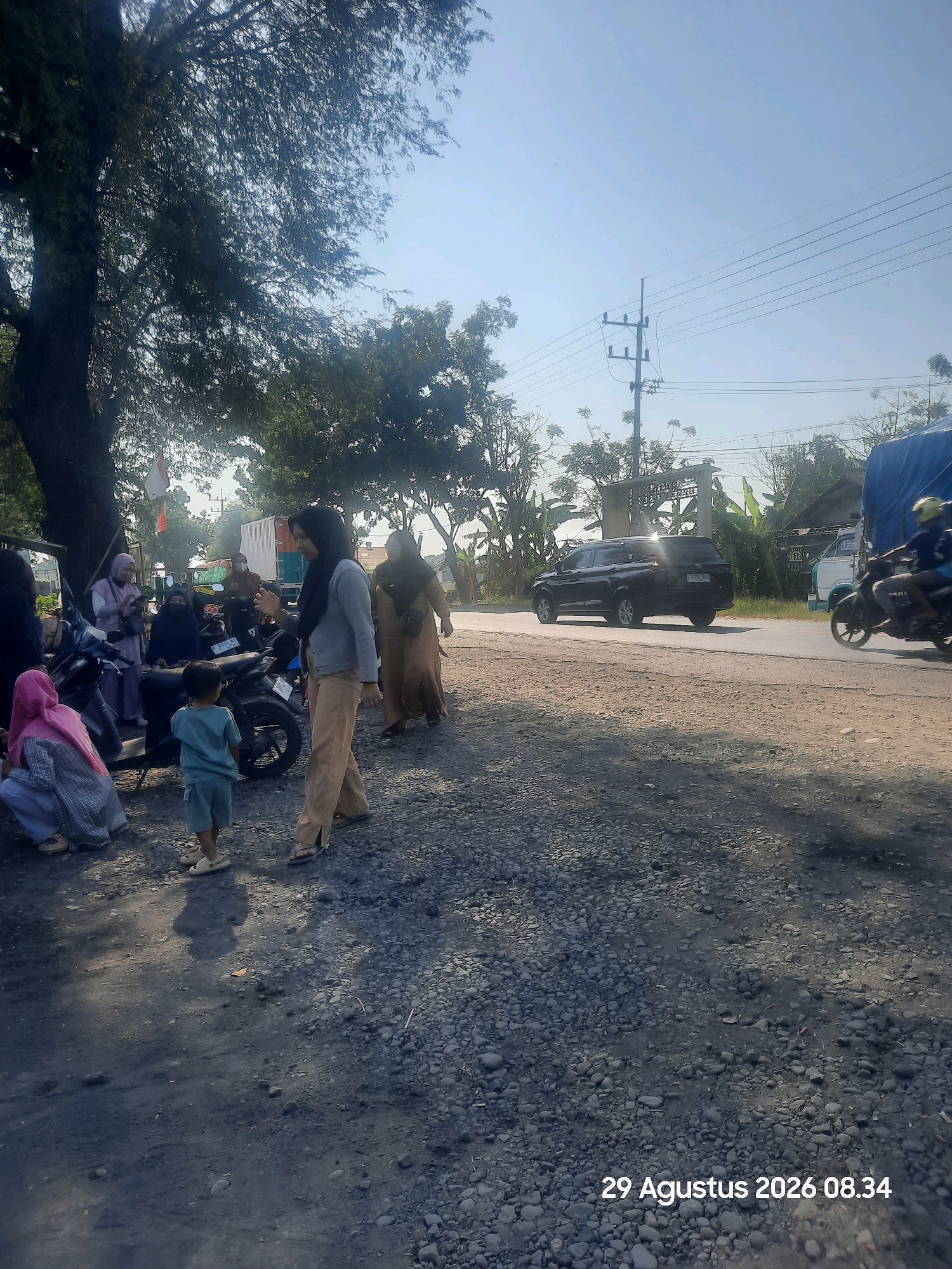 An outdoor scene in Sukokerto, Indonesia, captures a group of people and vehicles on a road with a gravel shoulder under a clear sky. A dark-colored SUV is driving on the paved road, while a motorcycle with two riders is also in motion. Several individuals are standing or sitting on the gravel shoulder near parked motorcycles. One person is wearing a light blue shirt and shorts, and others are dressed in traditional Indonesian attire, including hijabs. A large tree with dense foliage shades a portion of the scene. In the background, a white truck and a structure with a sign are visible. The date stamp "29 Agustus 2026 08.34" is present in the lower right corner.