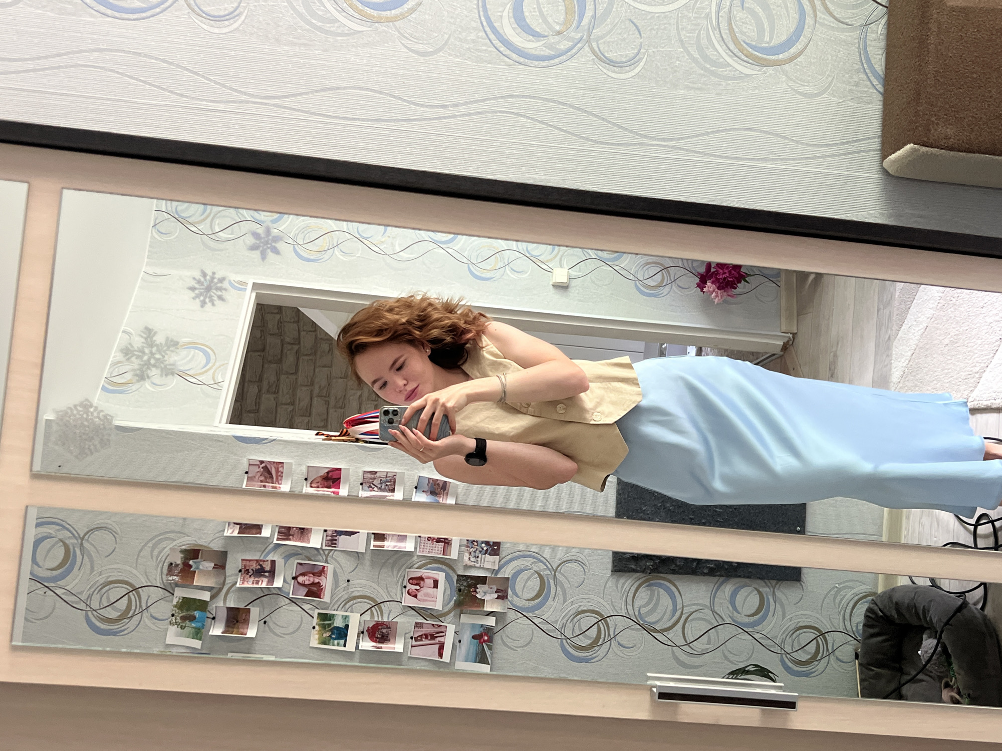 In the city of Chaikovsky, Russia, a young woman with curly red hair takes a selfie in a full-length mirror. She is wearing a light beige sleeveless vest over a light blue satin skirt. The mirror reflects a wall with light blue and grey swirling patterns, interspersed with snowflake decorations. On the left side of the mirror, a collection of small photographs is pinned to the wall, depicting various scenes and people. In the reflection, the woman is holding a smartphone in her right hand and is wearing a black watch on her left wrist and bracelets on her right wrist. A doorway is visible in the background reflection, leading to another room. To the left of the mirror, a dark grey pet bed is visible, and further to the left, a closet door with a vertical metallic handle.
