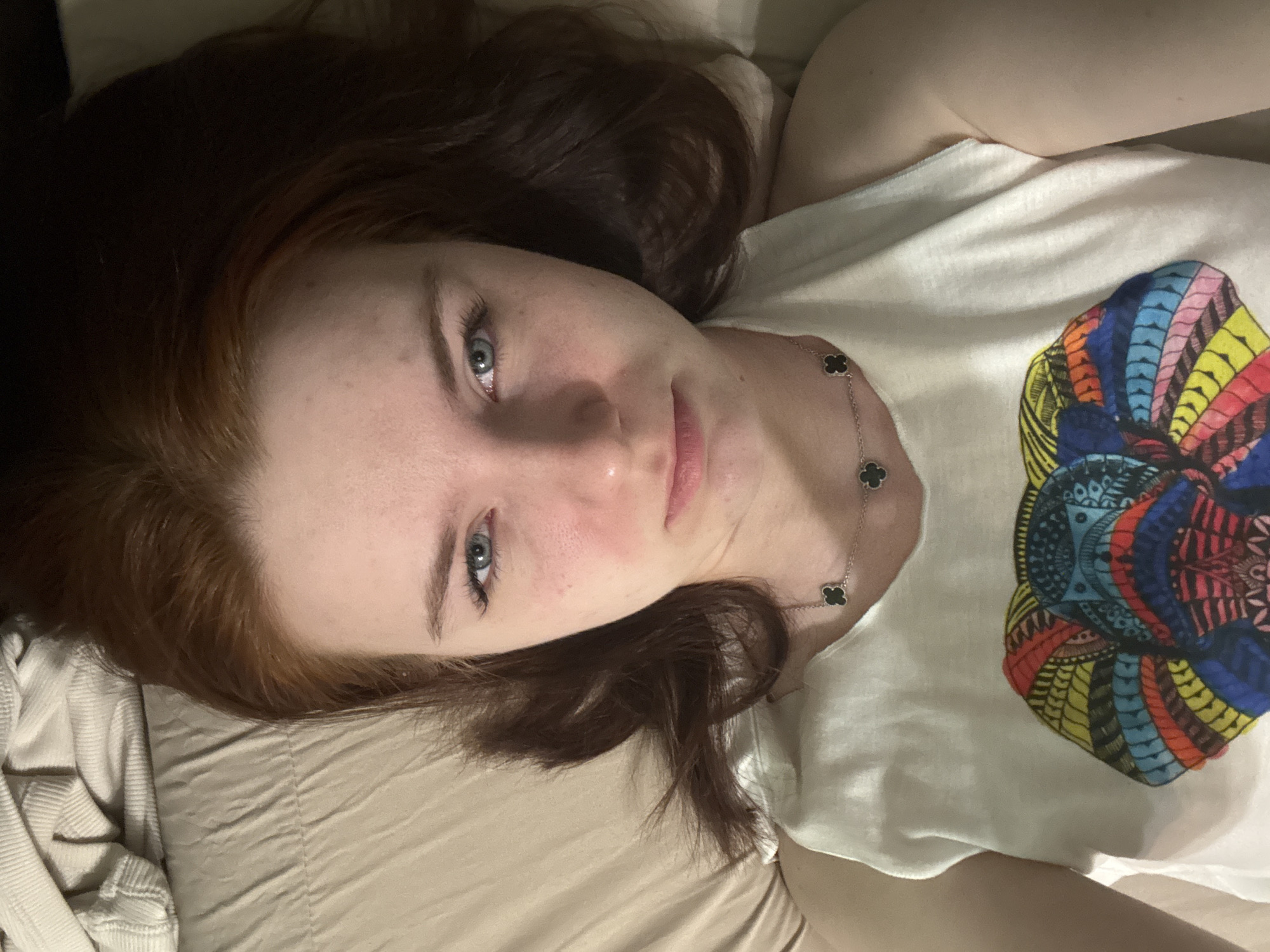 A woman lies on a bed in an unknown location. She has fair skin, blue eyes, and auburn hair that transitions to a darker brown. She wears a white t-shirt with a colorful circular graphic on the right side. She also wears a silver chain with four-leaf clover shaped pendants. The bed has beige bedding.