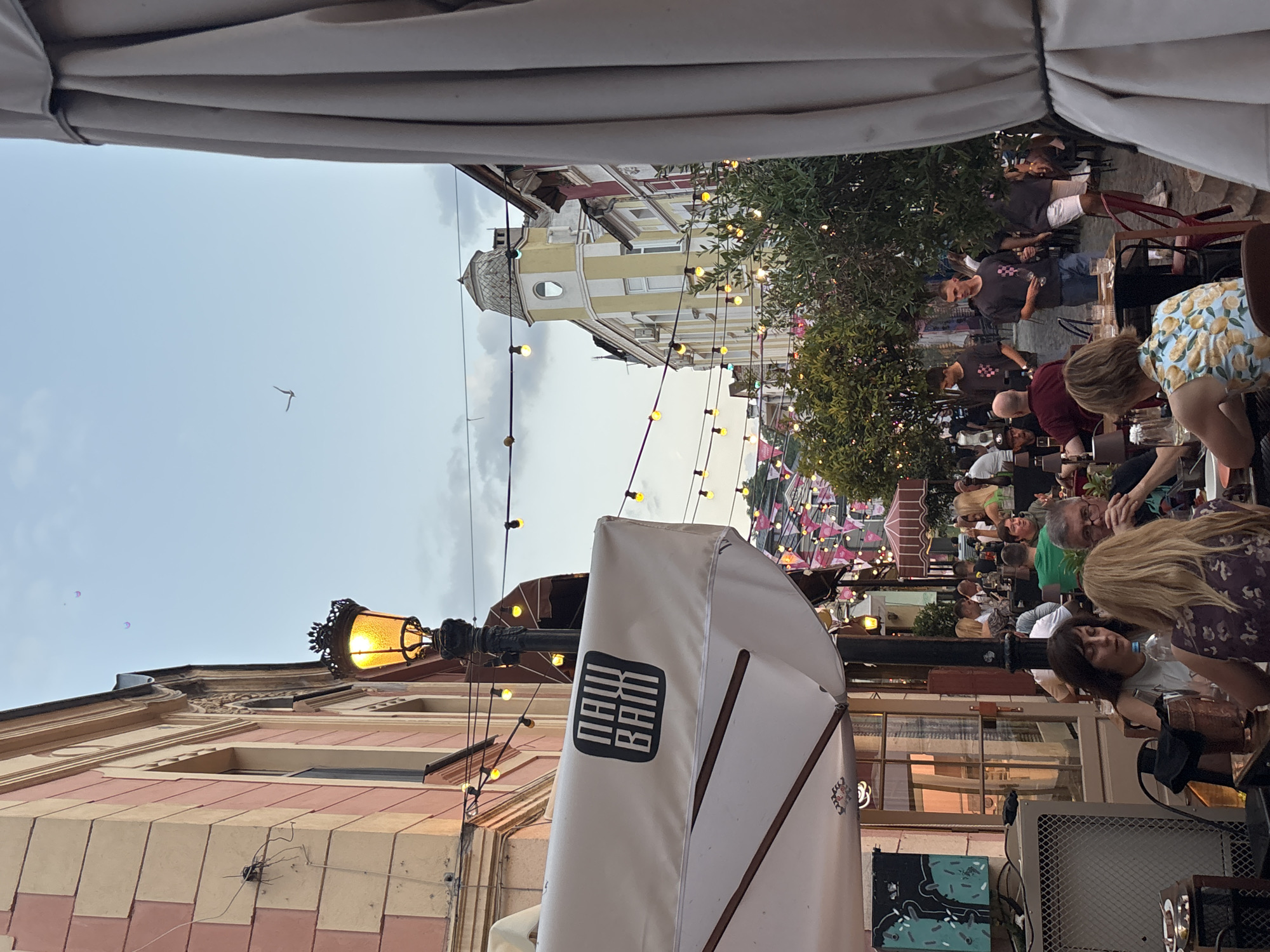 This image depicts an outdoor seating area at a cafe or restaurant, likely in a European city given the architectural style and string lights. The time of day appears to be late afternoon or early evening, as indicated by the soft, diffused lighting and the glow of string lights. The weather seems pleasant, with a clear sky.
The foreground is dominated by a white umbrella, partially obscuring the view, with a black logo featuring white vertical lines and the text "NORA" above "BRAT" in a stylized font. To the left, a building with a pink and cream facade is visible, featuring decorative cornices and windows. A vintage-style lamppost with an illuminated amber light stands prominently.
In the background, a bustling scene unfolds. Numerous people are seated at tables, engaged in conversation and enjoying themselves. Some appear to be tourists or locals relaxing. Behind the patrons, more buildings with yellow and white striped facades and a tower with a circular window are visible, adorned with festive string lights that contribute to a warm and inviting atmosphere. Trees with lush green foliage are interspersed throughout the scene. A single bird is captured mid-flight in the sky. The overall mood is relaxed and convivial, suggesting a pleasant social gathering.