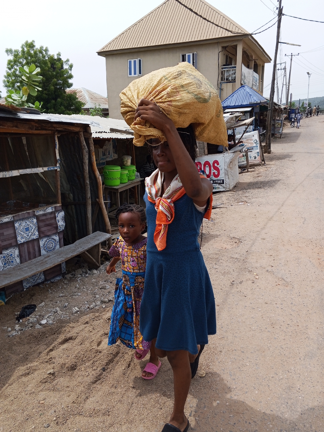 A woman and a young child walk along a dusty road in Adankolo, Nigeria. The woman, positioned prominently in the foreground, balances a large, light-colored sack on her head, supporting it with her right hand. She wears a knee-length blue dress, a patterned orange and white scarf around her neck, and eyeglasses. Her expression is neutral. Beside and slightly behind her, a small child looks towards the camera with a curious expression. The child is dressed in a brightly patterned traditional dress featuring yellow, orange, blue, and purple colors, and wears pink sandals.
The setting appears to be a semi-urban or rural street during the daytime, under what seems to be an overcast sky, as there are no harsh shadows. To the left of the subjects, several rustic market stalls or small shops with corrugated metal roofs and walls line the road. One stall has blue and white patterned tiles on its lower wall and a wooden bench. Green plastic containers are visible near another stall. In the mid-ground, a larger, two-story building with a light-colored facade, a beige roof, and blue-framed windows stands prominently. Utility poles and power lines run along the right side of the road, which stretches into the distance, revealing a slight uphill incline and distant hills.
The primary activity is the woman transporting goods on her head while walking with the child. On a white sign near the larger building, the red letters "POS" are visible, with "SERVICE HERE" written below. The ground is a mix of sand and dirt.