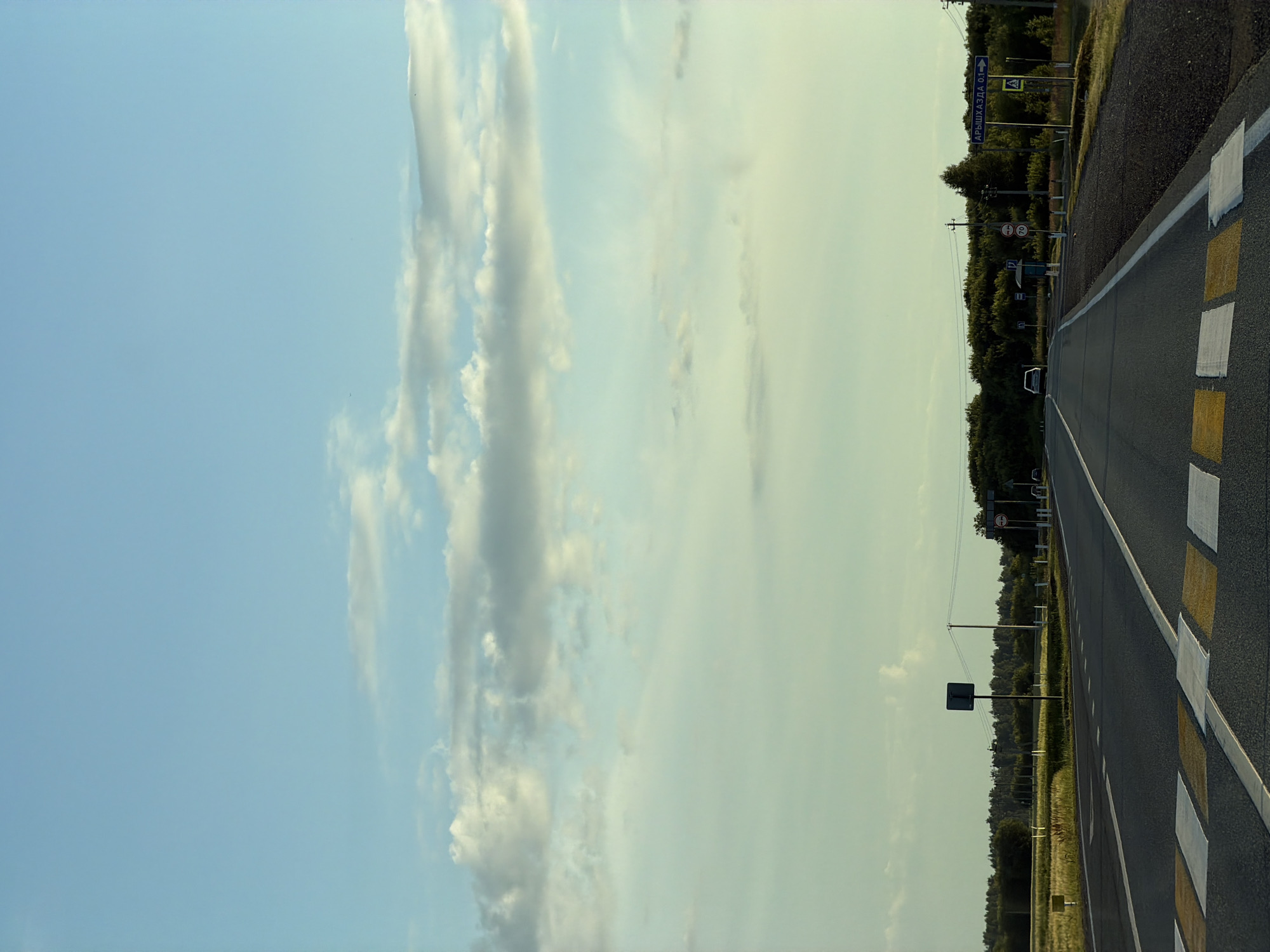 The image depicts a road in Aryshkhazda, Russia. The road is paved and has clear lane markings, including a pedestrian crossing. There are signs indicating speed limits and directions to Aryshkhazda, with a distance of 0.1 kilometers. A white car is visible on the road, and trees and fields line the sides. The sky is partly cloudy, and the overall lighting suggests it is either early morning or late afternoon.