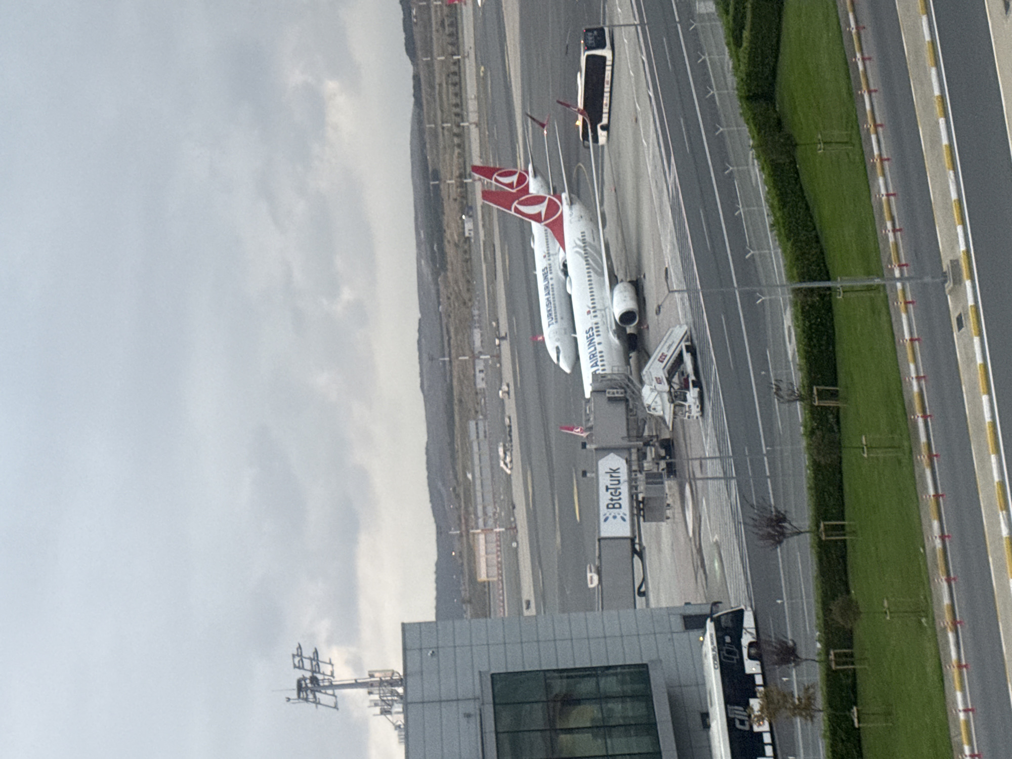 This is an exterior, medium-range, eye-level shot of Istanbul Airport in Arnavutköy, Turkey. The scene is dominated by an overcast sky. In the foreground, the side of a modern airport terminal building, constructed of gray panels and featuring large glass windows, is visible on the left. A jet bridge with "BteTurk" branding extends from the terminal towards two parked Turkish Airlines airplanes. The airplanes are white with red Turkish Airlines logos on their tails. A passenger bus, branded "CİB BUS," is positioned on the tarmac to the left of the terminal building. In the middle ground, a yellow and white striped bus is parked near the jet bridge. Further back, more airport infrastructure, including runways and other buildings, can be seen, with a gently rolling, tree-covered landscape in the background. The overall atmosphere is one of activity at a major international airport under cloudy weather conditions.