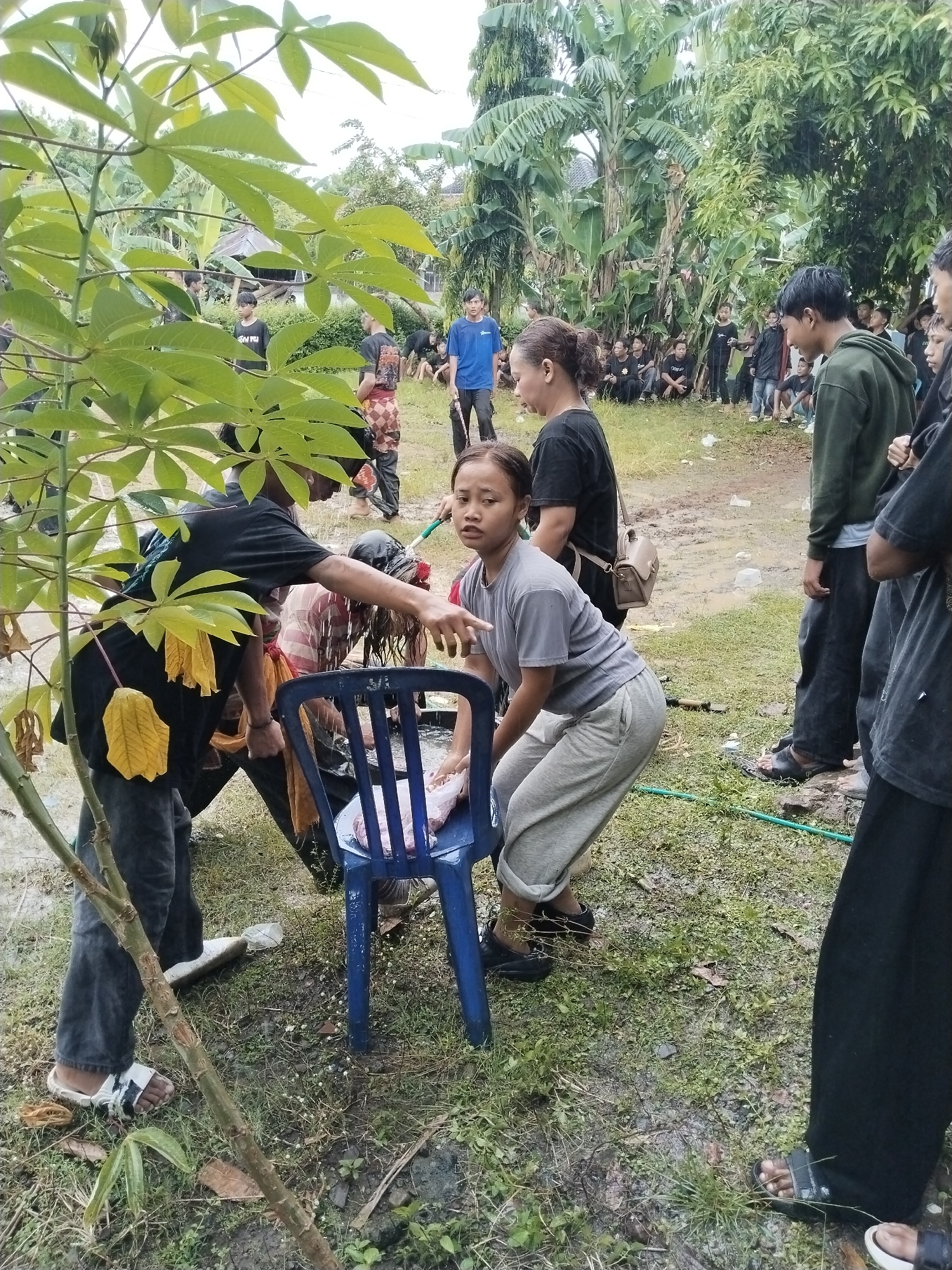 The image shows a group of people gathered outdoors in what appears to be a grassy area, possibly in Karang Tengah, Indonesia. The ground is wet and muddy, suggesting recent rain. In the foreground, a woman wearing a gray shirt and gray pants is crouching next to a blue plastic chair. Another individual, wearing a black shirt with yellow accents, is interacting with someone seated in the chair, possibly performing some sort of ritual or washing. There is what appears to be meat on the seat. Behind the main group, there is a crowd of people watching. Some are standing, while others are seated on the ground. The background shows dense foliage and trees.