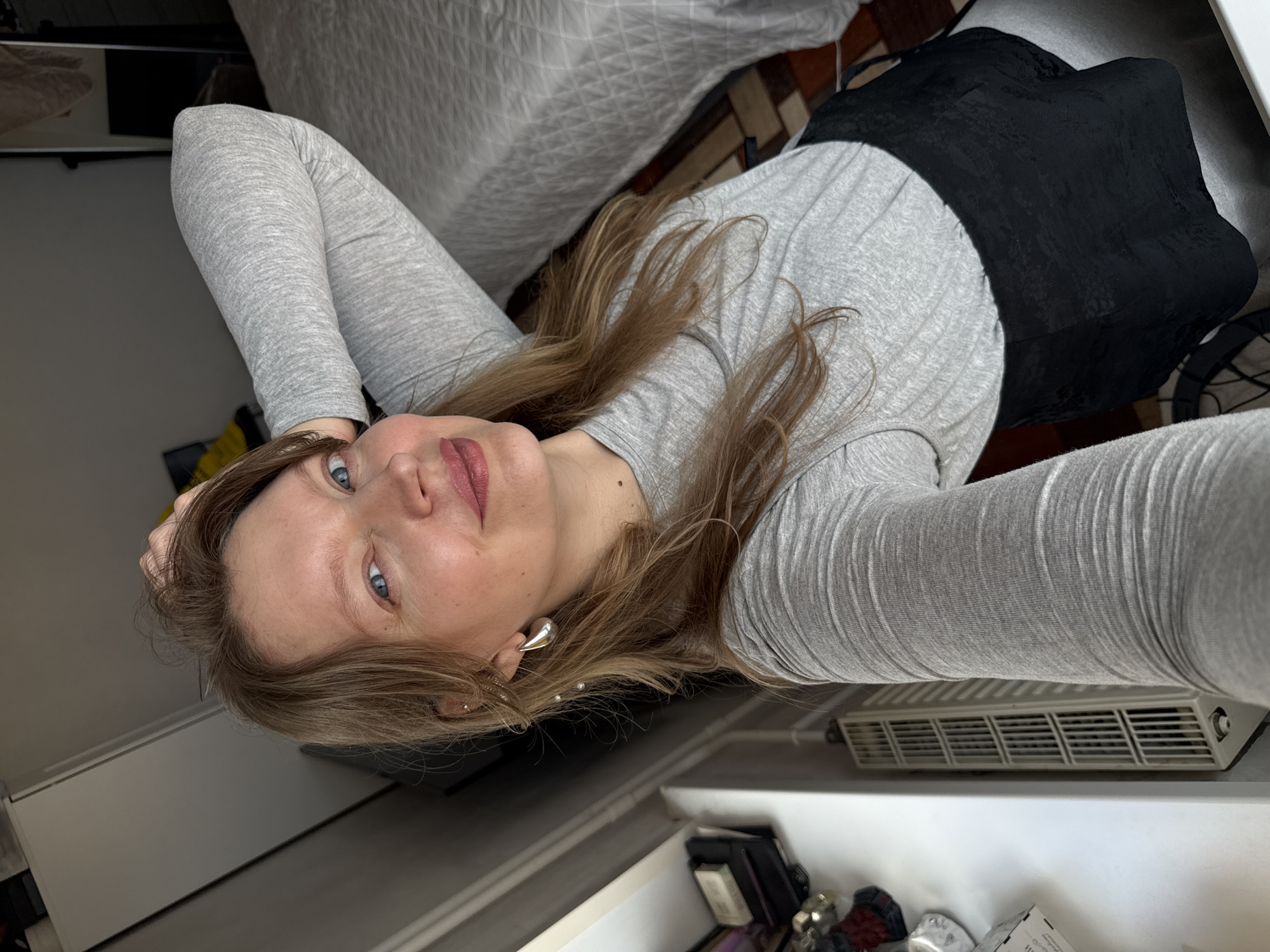 A woman with blonde hair and blue eyes is taking a selfie in what appears to be a bedroom in Izhevsk, Russia. She is wearing a grey long-sleeved shirt and a black corset. She has her right arm raised behind her head, and her left arm is not visible. The room is furnished with a bed, a radiator, and a wardrobe. The lighting suggests it is daytime.