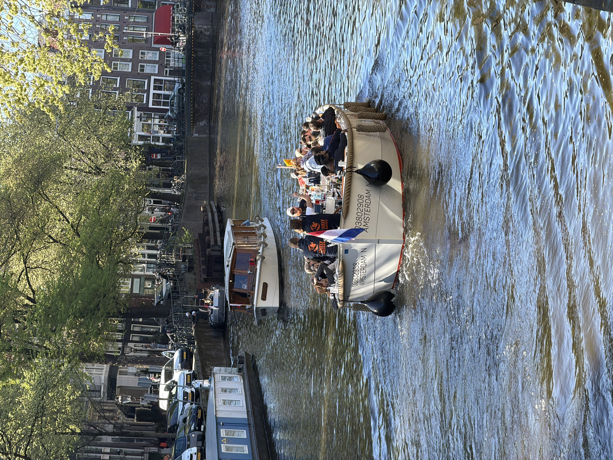 The image, if oriented correctly in landscape, captures a vibrant canal scene in Amsterdam on a bright, sunny day. The blue-green water of the canal occupies the central and right portions of the frame, showing ripples and reflections of sunlight.
In the foreground, a white tour boat is prominently featured, packed with numerous passengers enjoying a leisurely cruise. The boat is moving, creating a small wake behind it. On its hull, the text "08802908 AMSTERDAM N" is clearly visible, along with the branding "LOVELY CRUISES". A person wearing a black t-shirt with an orange insect-like logo and the "LOVELY CRUISES" text is standing towards the front of the boat. A small Dutch flag is attached to the boat's side. Black fenders hang from the boat's front.
Further back on the canal, to the left of the main tour boat, a second, smaller boat with a wooden cabin and fewer occupants is also navigating the water.
The left bank of the canal is lined with lush trees featuring light green, fresh foliage, indicating spring or early summer. Behind the trees, characteristic Amsterdam canal houses with dark brick facades, white-trimmed windows, and red awnings are visible. The street along the canal bank is populated with numerous parked bicycles, parked cars, and a red circular "no entry" sign.
The scene conveys a relaxed, tourist-friendly atmosphere, with clear skies and ample sunshine.