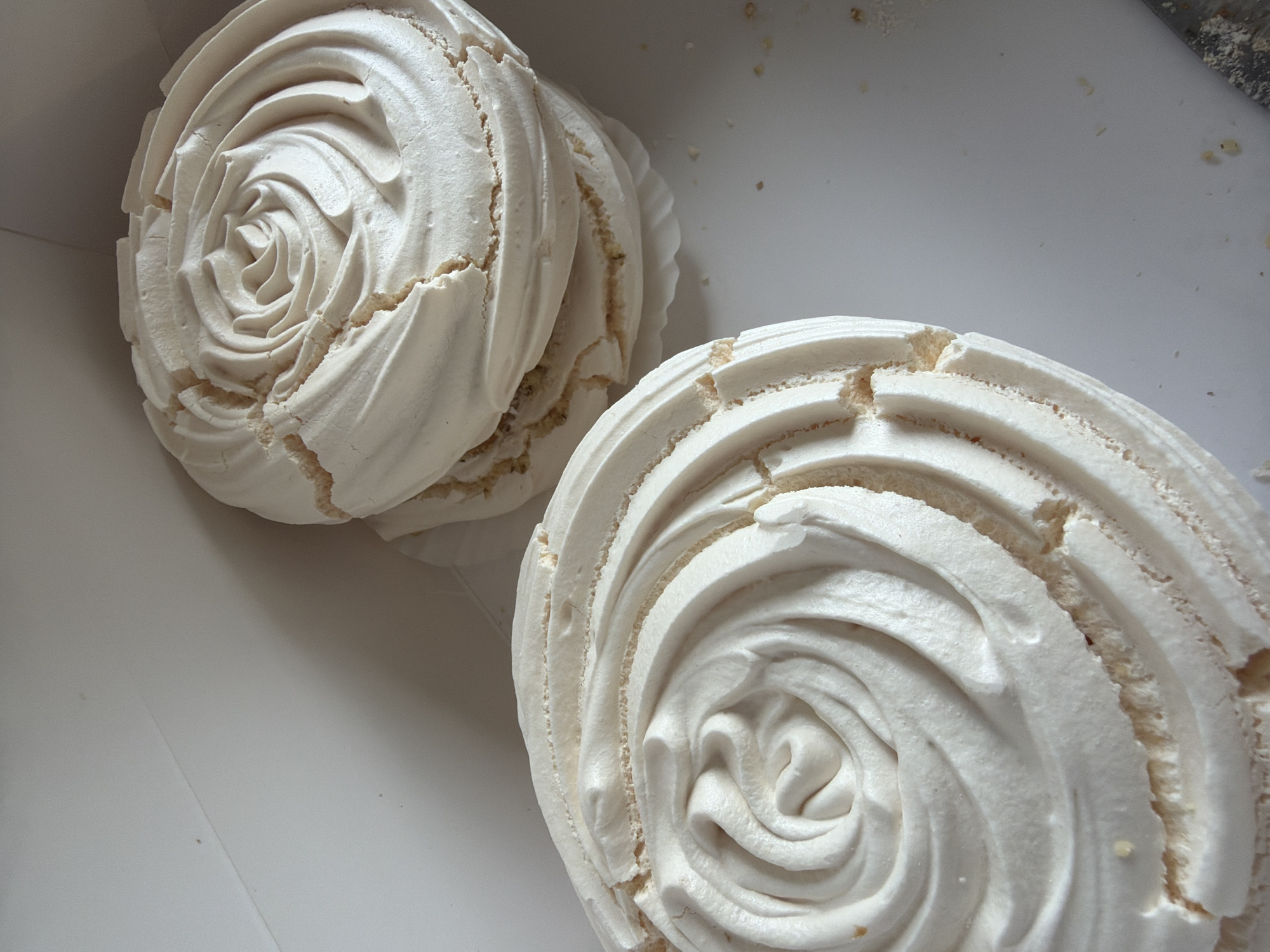 The image captures a close-up view of several delicate meringue shells, likely prepared for desserts, presented in a clean, white container. The scene is set indoors, with the location context being Yerevan, Armenia.
The primary subjects are the meringues themselves, characterized by their intricate, swirled piping pattern that resembles a rose or a tight spiral. They are predominantly off-white or cream-colored, suggesting a perfectly baked consistency. The surface of the meringues shows minor cracks and fissures, revealing a slightly rougher, more porous texture beneath the smooth piped exterior, which is typical for baked meringues.
In the foreground, one large meringue is prominently displayed, its detailed swirls and delicate cracks clearly visible. Behind it, at least two more meringues are partially visible, stacked or overlapping, suggesting a batch of these confections. The background consists of the stark white interior of the container, with subtle shadows defining its edges and occasionally showing tiny crumbs or specks, possibly from the meringues themselves.
The image focuses purely on the food items, with no people, discernible activities, or visible text. The lighting is soft and even, highlighting the textures and the elegant form of the meringues without harsh shadows. The overall impression is one of simple elegance and fresh, homemade confectionery.