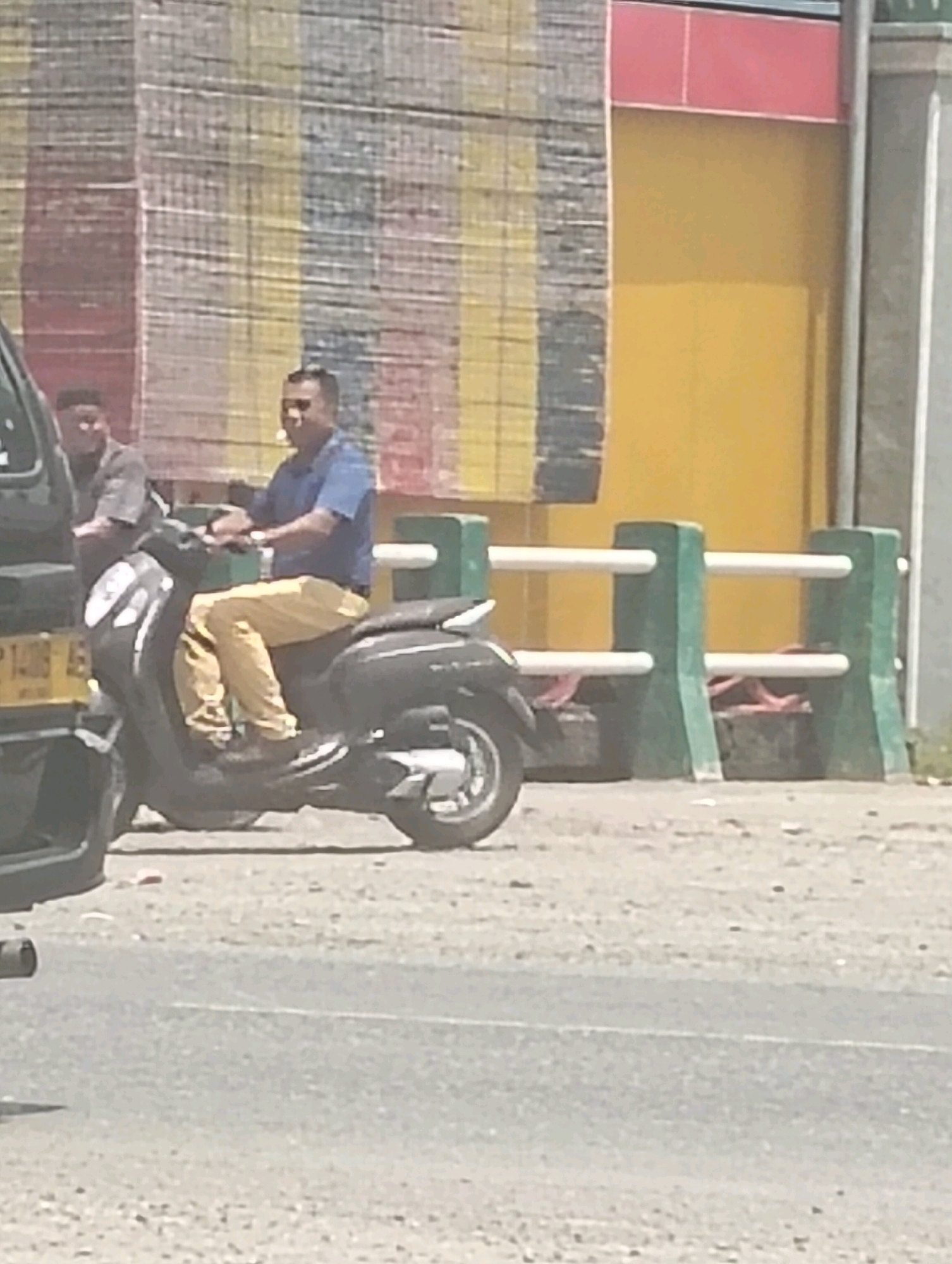 A man wearing a blue shirt and yellow pants is riding a black scooter on a paved road. Another person, partially visible, is seated on a dark-colored vehicle behind him. The background features a building with a yellow wall and a striped facade, along with a green and white railing. The scene appears to be outdoors, under daylight.
