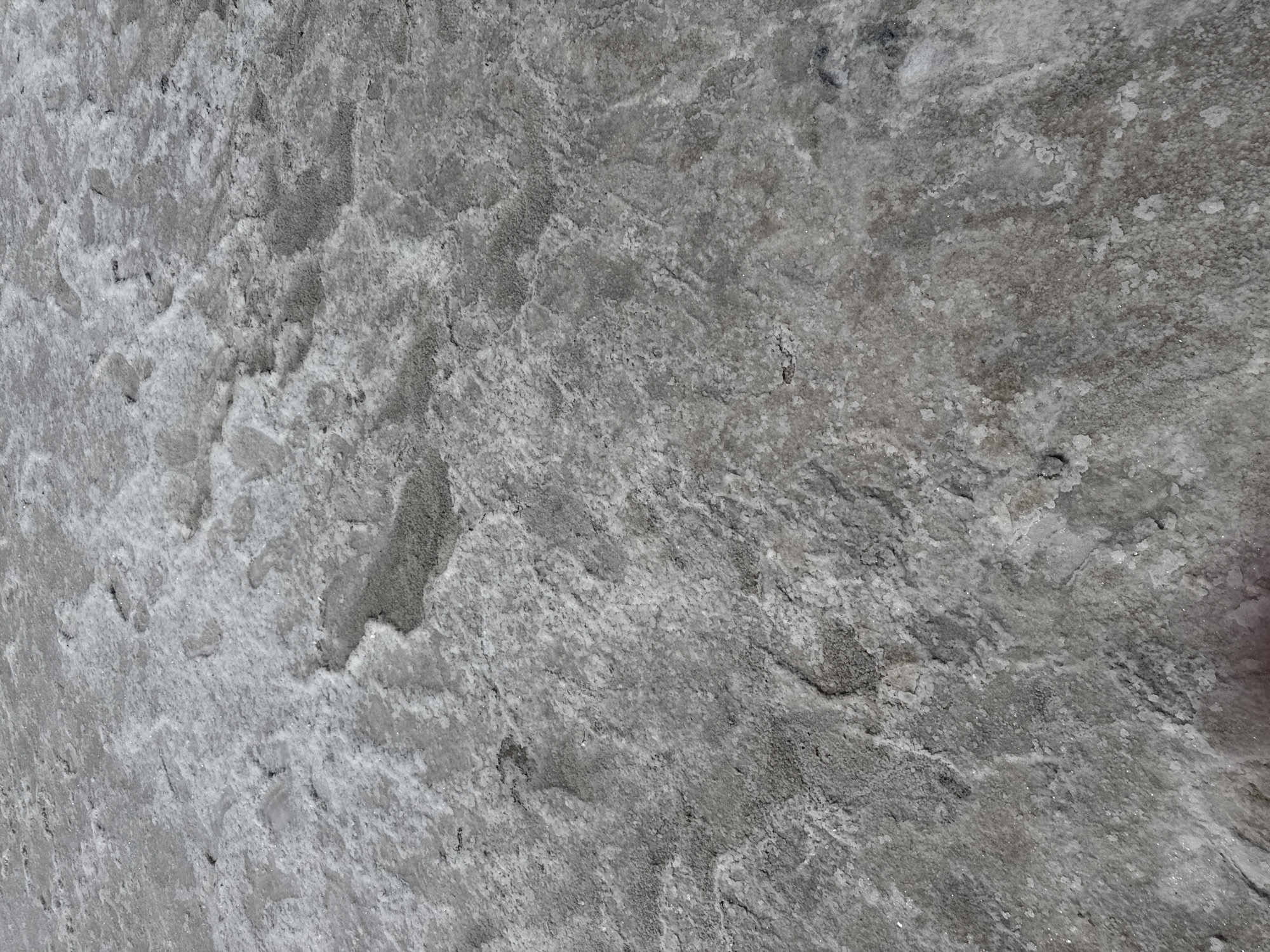 The image shows a close-up, overhead view of cracked, dry mud or salt flats with areas of hardened, crystalline deposits. The terrain is uneven, with some depressions and raised sections, creating a textured surface. Small pebbles and darker, granular patches are interspersed with the lighter, salt-like formations. The lighting suggests an outdoor environment, possibly under daylight, casting subtle shadows that emphasize the texture. The overall color palette is muted, dominated by shades of gray and white. There are no discernible people or specific objects that indicate a particular activity or event. The scene appears to be a natural landscape feature, likely an area that has recently experienced drying conditions, such as a dry lakebed or salt pan. The location is stated to be Bursol', Russia, which is known for its salt lakes and mineral deposits, fitting the visual evidence in the image.
