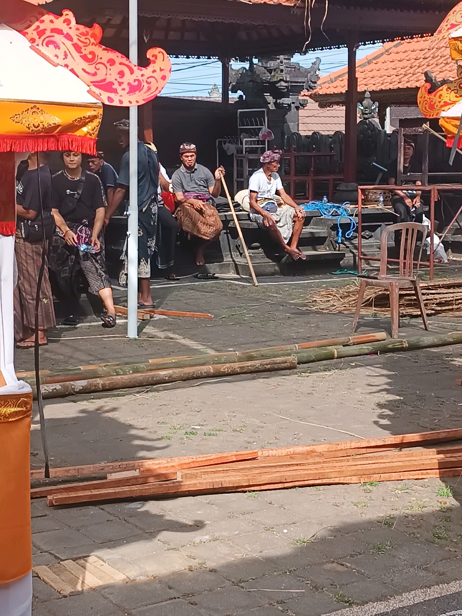 This image captures a scene outdoors in Selemadeg, Indonesia, likely during a cultural event or preparation. In the foreground, a stack of lumber lies on a paved surface, with several bamboo poles also visible. To the right, a plastic chair sits near a pile of dried grass or reeds.
Further into the scene, several men are present, some seated and others standing. Many are wearing traditional Balinese attire, including sarongs and headbands. The men appear to be engaged in observation or rest, with one man holding a wooden pole. Behind them, there is a building with a tiled roof and intricate architectural details, possibly a temple or traditional structure. The environment suggests a sunny day with distinct shadows cast by the structures and objects. Decorative elements, including ornate umbrellas or canopies, are visible at the top left and right edges of the image, adding to the festive or ceremonial atmosphere.