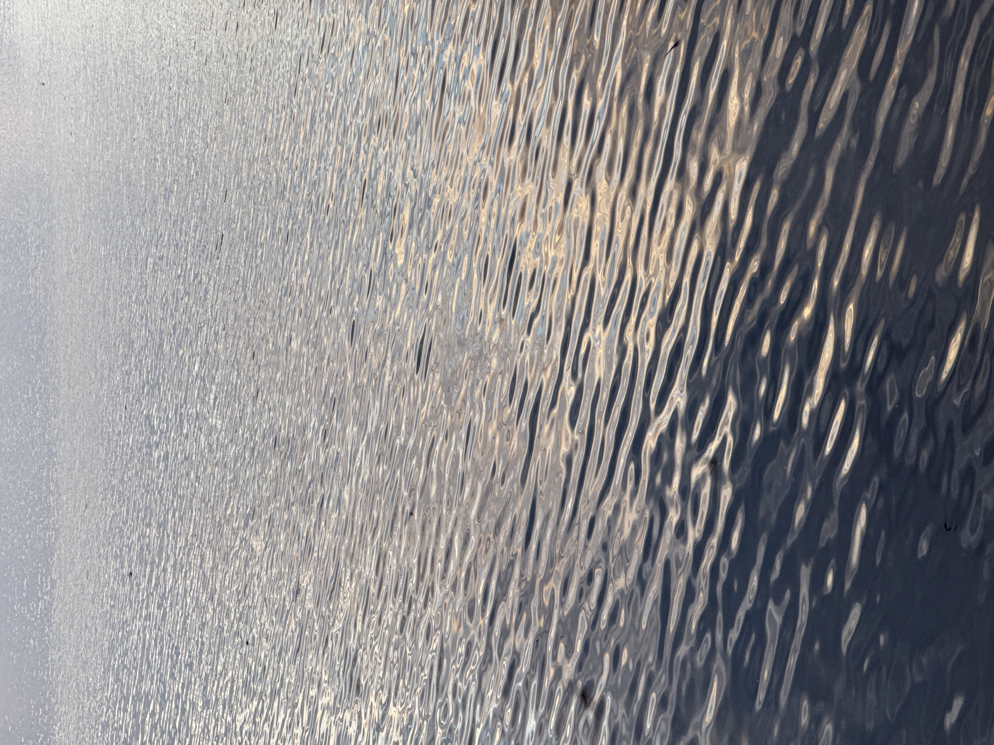 The image shows a close-up view of rippling water, likely a body of water in Chaikovsky, Russia, such as the Kama River or a local reservoir. The water surface reflects light, creating patterns of light and dark with horizontal waves. There are no visible people, structures, or specific landmarks in the frame. The overall impression is one of natural texture and light play on a water surface.