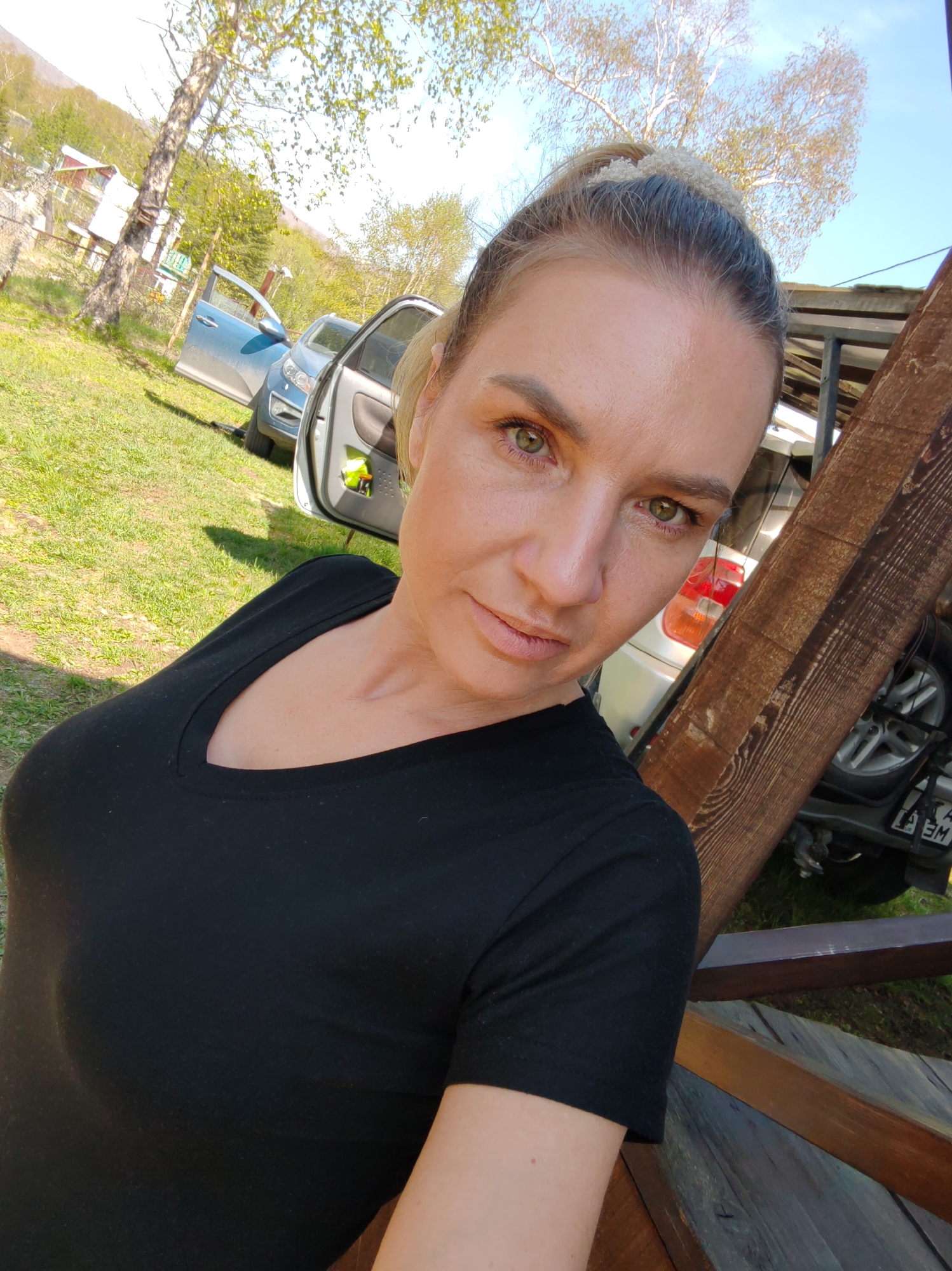 An outdoor, eye-level, slightly upward-angled portrait shot captures a light-skinned woman, likely in her 30s or 40s, looking directly at the camera with a neutral expression. Her blonde hair is pulled back, revealing darker roots. She has green eyes with subtle eye makeup and light-colored lips. She wears a black, short-sleeved, v-neck top. A distinctive wooden beam frames the right side of the image. The background reveals a sunny, green grassy area with several trees, one appearing to be a birch. Two cars are visible: a light blue SUV-style vehicle with its front driver's side door open, and a lighter-colored vehicle (possibly silver or white) further back, displaying a partial license plate with "A" and "M" visible. Distant hills or mountains are present, and a building with a red roof is discernible on the left. The scene suggests a rural or semi-rural location in Unknown, Unknown.