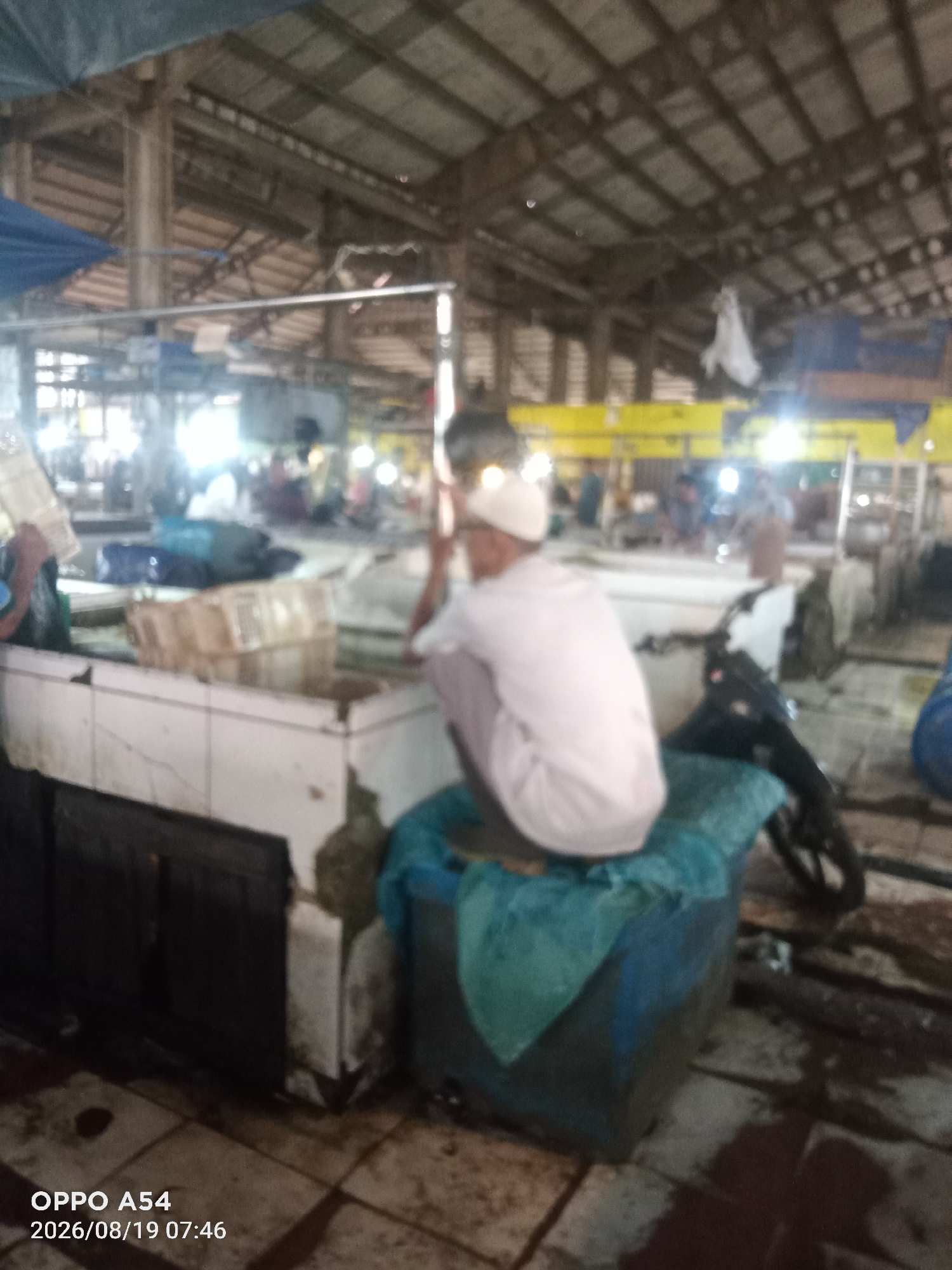 The image depicts a blurry scene within an indoor market, likely in Jambi, Indonesia, given the timestamp indicating a 2026 date and an Indonesian phone model. A person wearing a white cap and light-colored clothing is seen squatting on a blue container, facing away from the camera. The environment appears to be a stall area with white tiled counters and wooden structures overhead. There are various objects, including crates, tarps, and a motorcycle parked in the background. The lighting suggests it is daytime, with some artificial lights also present. The overall atmosphere is that of a busy marketplace, though the low resolution and blurriness obscure finer details and the specific location within Jambi.