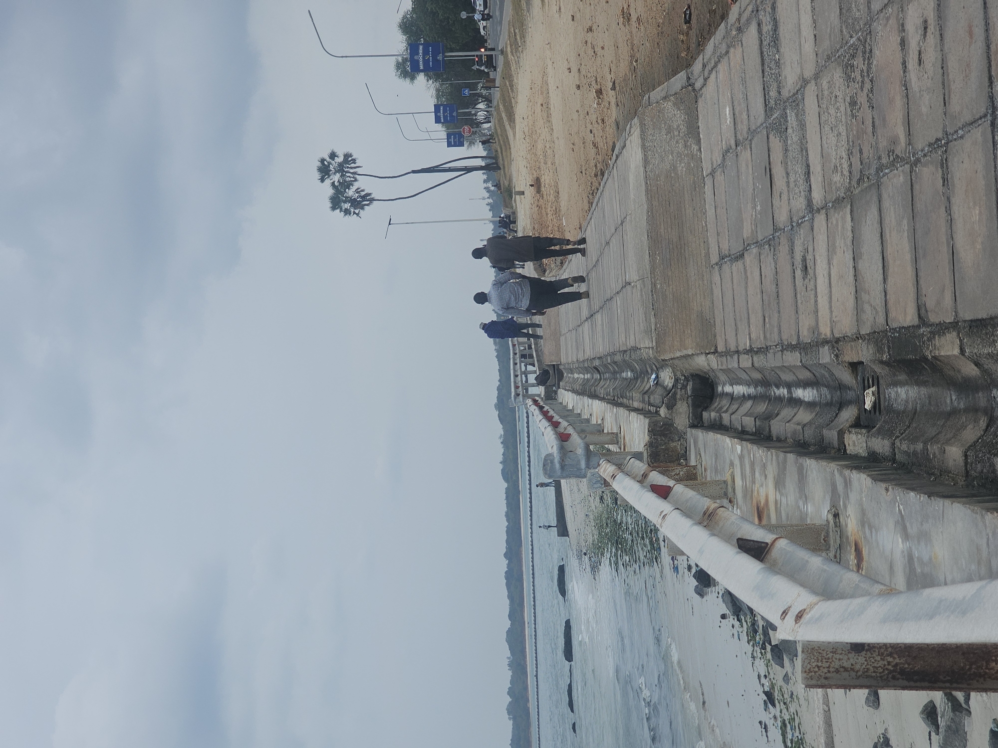A paved walkway runs along a beach on the coast of Dar es Salaam, Tanzania, with a guardrail separating it from the sea. Two men in casual attire are walking away from the camera on the walkway. The beach is sandy and scattered with rocks and some debris. Palm trees are visible in the distance, along with streetlights and a road with a few vehicles. The sky is overcast and the sea appears calm with gentle waves.