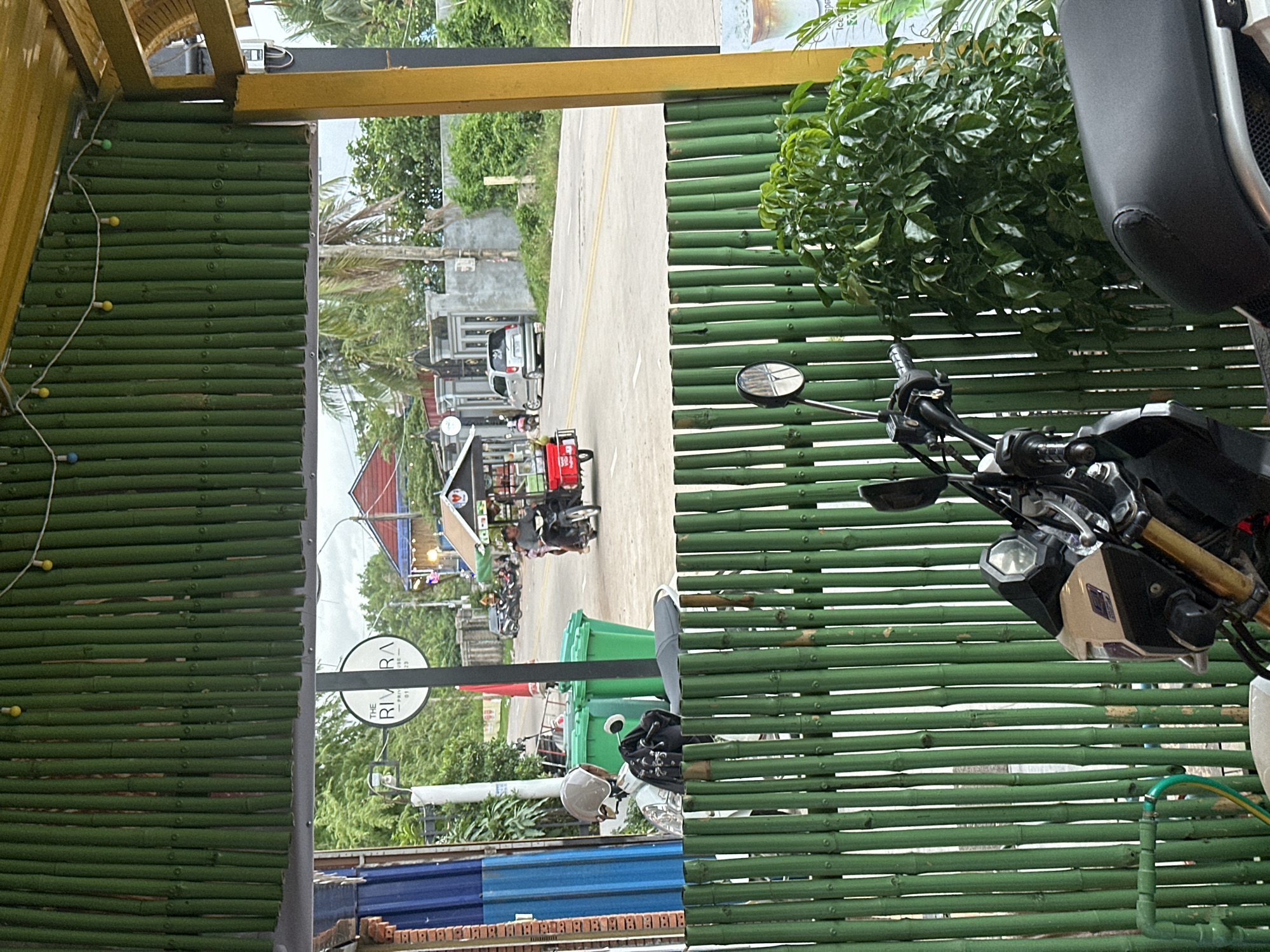 The image depicts a street scene in Kampot, Cambodia, viewed from behind a green bamboo fence. In the foreground, the handlebars and front of a motorcycle are visible, along with a portion of its seat. Behind the fence, two large green trash bins stand next to a white motorcycle with a helmet and bag. Across the street, a silver car is parked, and several motorcycles are lined up. A man is riding a red three-wheeled vehicle, similar to a tuk-tuk, down the road. In the background, buildings with red roofs and other structures are visible, surrounded by lush green trees and palm trees. A sign in the distance reads "THE RIVIERA." The overall environment suggests a tropical, urban setting.