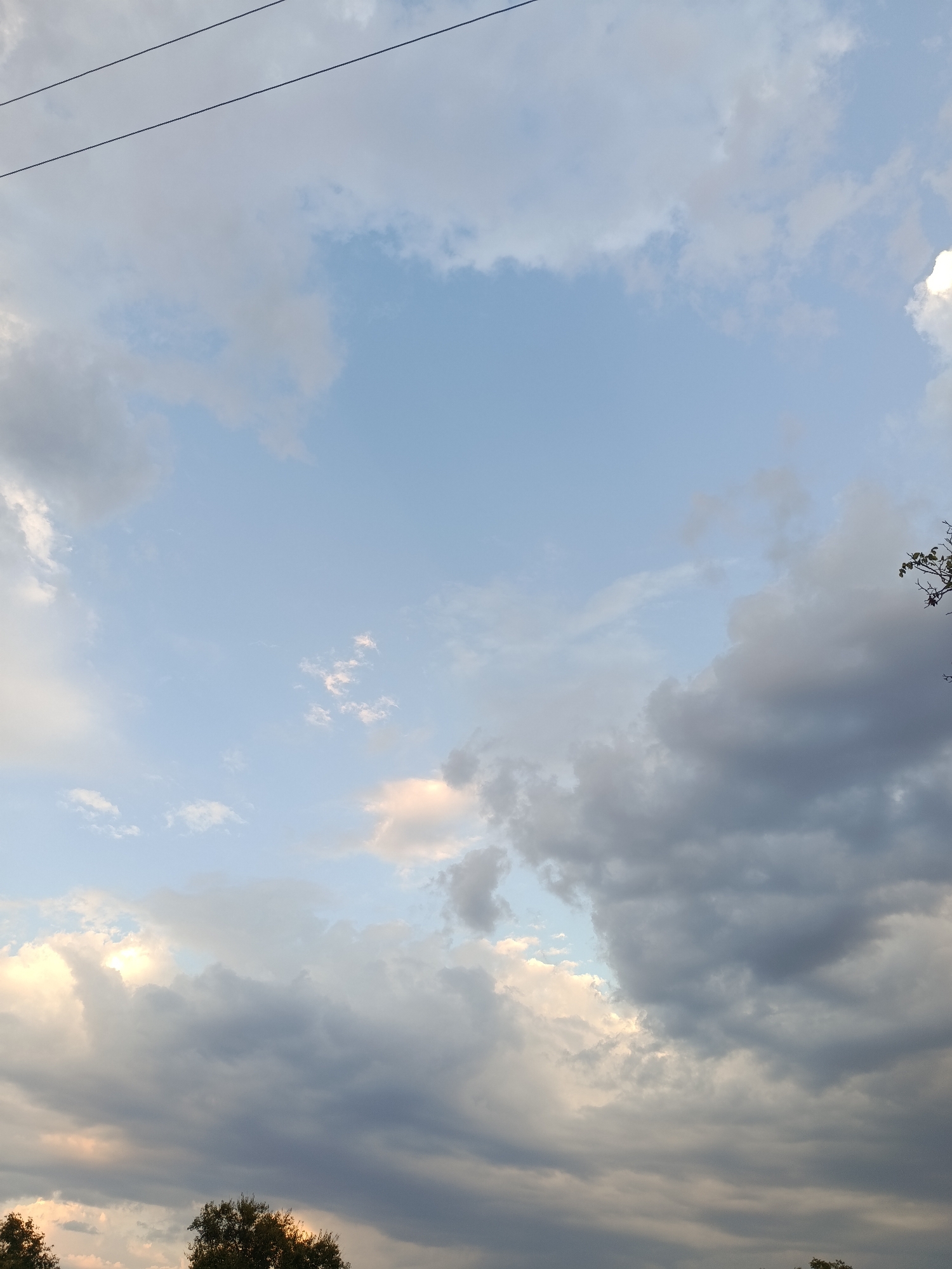 The image captures a cloudy sky with a clear patch of blue sky towards the upper center. The clouds are varied, with some appearing white and illuminated, while others are darker and more ominous. The bottom of the image shows the tops of trees, suggesting an outdoor scene. Two thin, dark lines, possibly power lines, are visible in the upper portion of the sky. The scene appears to be an overcast day in Smila, Ukraine. There are no people or specific actions depicted in the image.