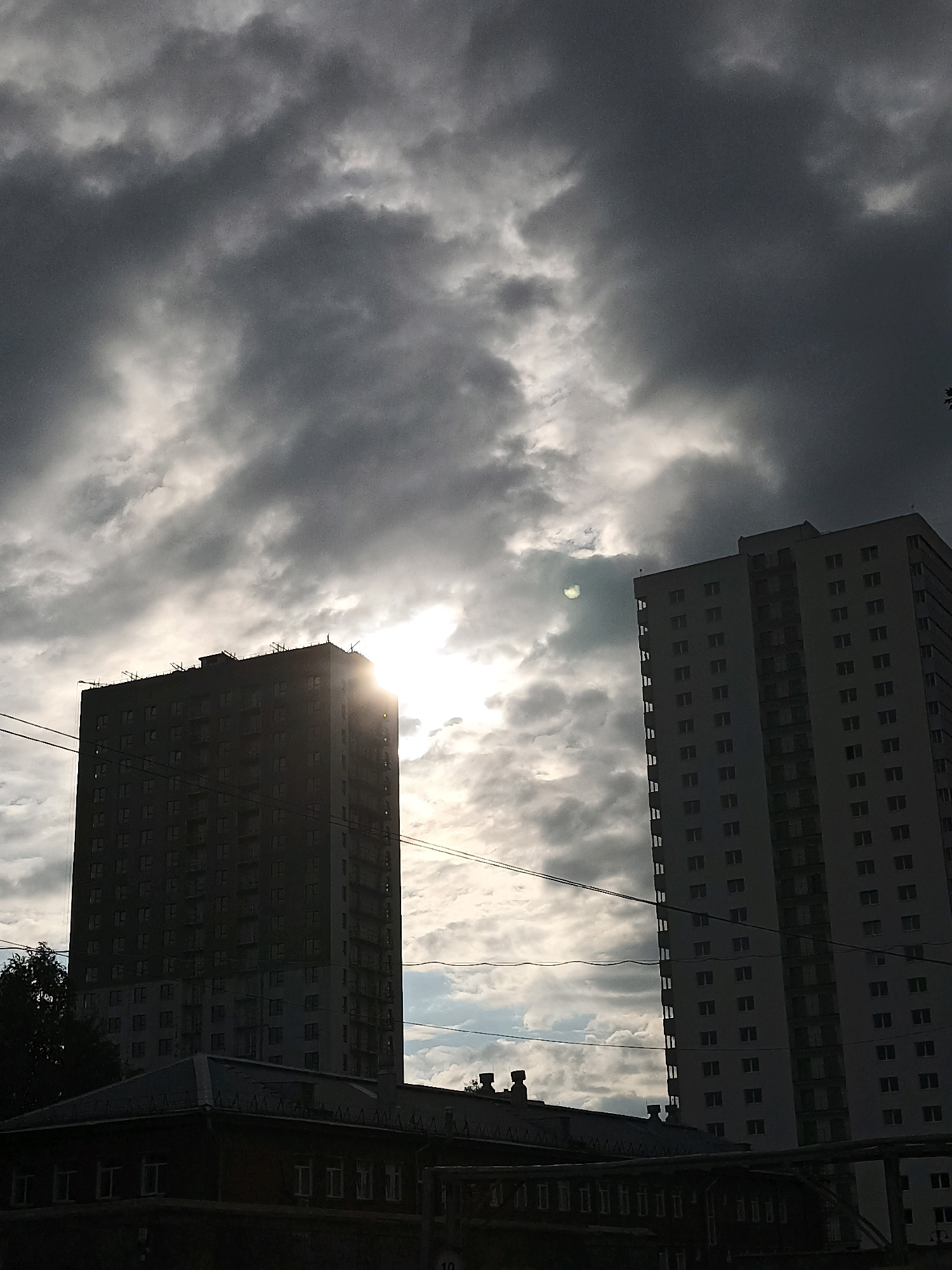 The image depicts a silhouetted view of two high-rise apartment buildings against a dramatic, cloudy sky. The sun is partially obscured by clouds, creating bright shafts of light. The building on the left appears to be under construction, with visible scaffolding on its upper floors. The building on the right is complete and shows a uniform pattern of windows. In the foreground, the dark silhouette of a lower-rise building with a gabled roof is visible. Overhead, several power lines cross the frame. The scene is overcast, with dark, heavy clouds dominating the upper portion of the image, contrasted by patches of sunlight. The overall impression is one of an urban landscape under a brooding sky.