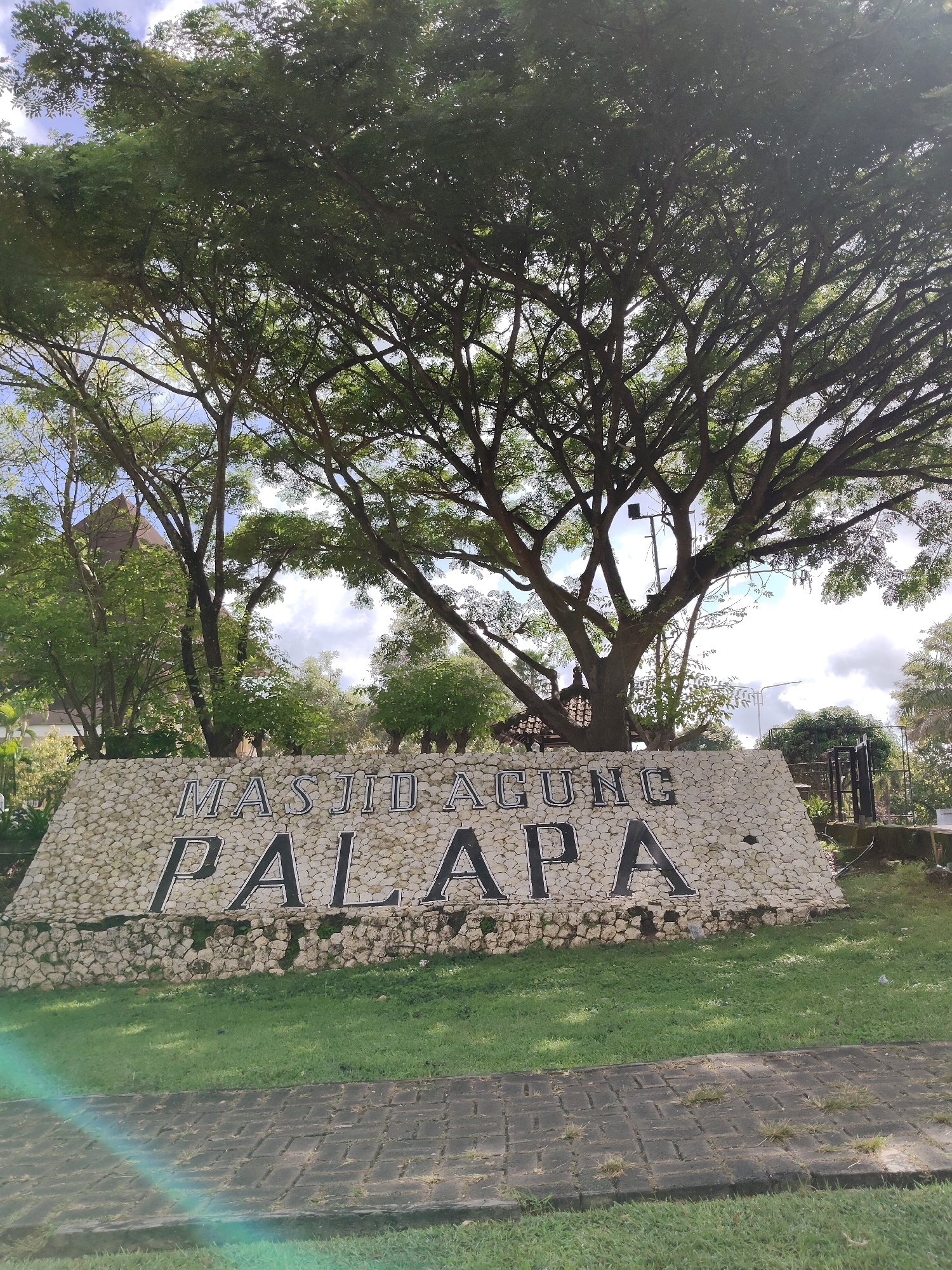 The image shows the signage for Masjid Agung Palapa in Ungasan, Indonesia. The sign is constructed from white stones with black lettering that spells out the name of the mosque. A large tree with dense foliage partially obscures the sign from view. Below the sign, there is a well-manicured lawn, and in the foreground, a paved pathway. The sky is visible through the branches of the tree, with some clouds present.