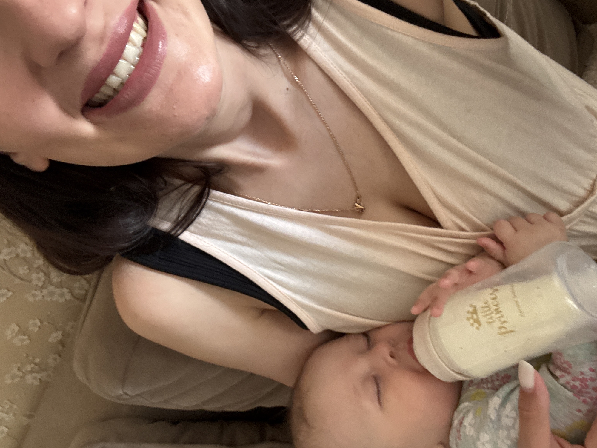 A woman lies on a soft surface, possibly a couch or bed, and smiles while holding a baby who drinks from a bottle labeled "Little Princess." The woman has dark hair and wears a beige top with a black undershirt. She also wears a delicate gold necklace. The baby, dressed in a floral-patterned outfit, has closed eyes and is focused on drinking. The background suggests an indoor setting. There is no information to indicate the location is in Sochi, Russia.
