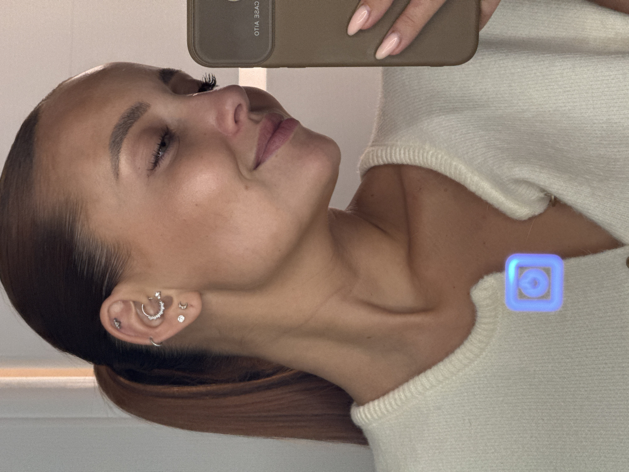 A close-up, self-portrait photograph depicts a woman's left profile, with her head slightly tilted down and a subtle smile. She has dark brown hair pulled back in a ponytail. Her left ear displays multiple piercings: a small stud in the tragus, a large crescent-shaped hoop adorned with clear stones in the conch, two small hoops and a stud in the helix region, and three studs (one small, one larger with a clear stone, and one shaped like a crescent moon) along the earlobe. Her skin appears clear, and her eyebrows are well-defined. Her eyelashes are prominent, suggesting mascara or extensions.
She is wearing a cream or off-white knitted sweater or cardigan with visible ribbing at the neckline. On her chest, below the neckline, a digitally superimposed, glowing blue square icon with a circular center design is visible. Her right hand, with neatly manicured, light pink/nude oval-shaped nails, holds a dark, possibly olive-green, smartphone case. The case has mirrored text, "OTIA 3SAD," visible on its surface, and part of the phone's camera lens is reflected. The background is indistinct and light-colored, suggesting an indoor setting. The image provides no identifiable geographical markers to determine its specific location, including the city of Chaikovsky, Russia.