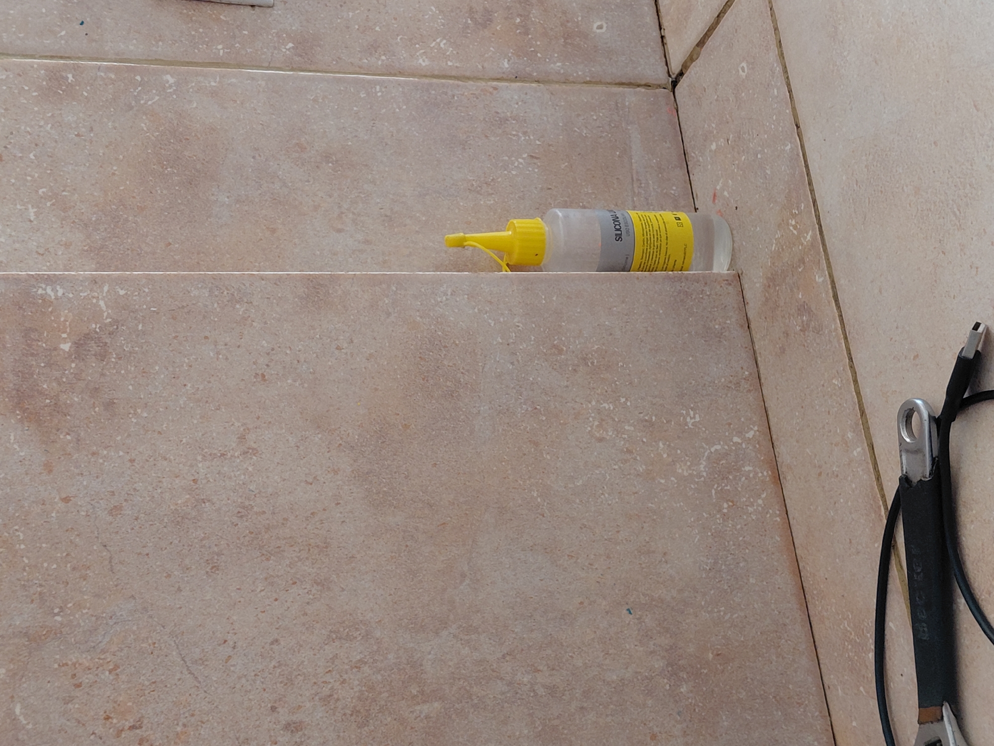 The image displays a close-up, slightly elevated view of a tiled surface, likely a floor or a step, in what appears to be an indoor or semi-enclosed setting in Santa Ana, Venezuela. The tiles are rectangular, predominantly light beige or tan, and feature a speckled, mottled pattern, suggesting a natural stone appearance or a ceramic mimic. Distinct grout lines, some showing minor imperfections or previous work, separate the individual tiles.
A small, clear plastic bottle with a bright yellow conical nozzle and cap is positioned horizontally on a grout line in the upper center of the frame. This bottle is clearly a sealant. Its yellow label features black text, with "SILICON" prominently visible, indicating it is a silicone product (likely "SILICONA" in Spanish). Additional smaller text on the label, possibly detailing product usage or type, is present but largely unreadable.
To the far right, partially visible at the edge of the frame, is a black cable or strap coiled around a metallic object. This object appears to be part of a hand tool, possibly a wrench or a similar utility implement, characterized by a silver-colored eyelet or loop at its end and a dark, possibly rubberized, handle.
There is no human activity depicted. However, the presence of the silicone sealant and the tool suggests that maintenance, repair, or construction work, such as sealing joints or applying caulk, has recently occurred or is underway. The lighting is neutral, consistent with an interior environment, providing no specific clues regarding the time of day or external weather conditions. The scene conveys a functional area within a building.