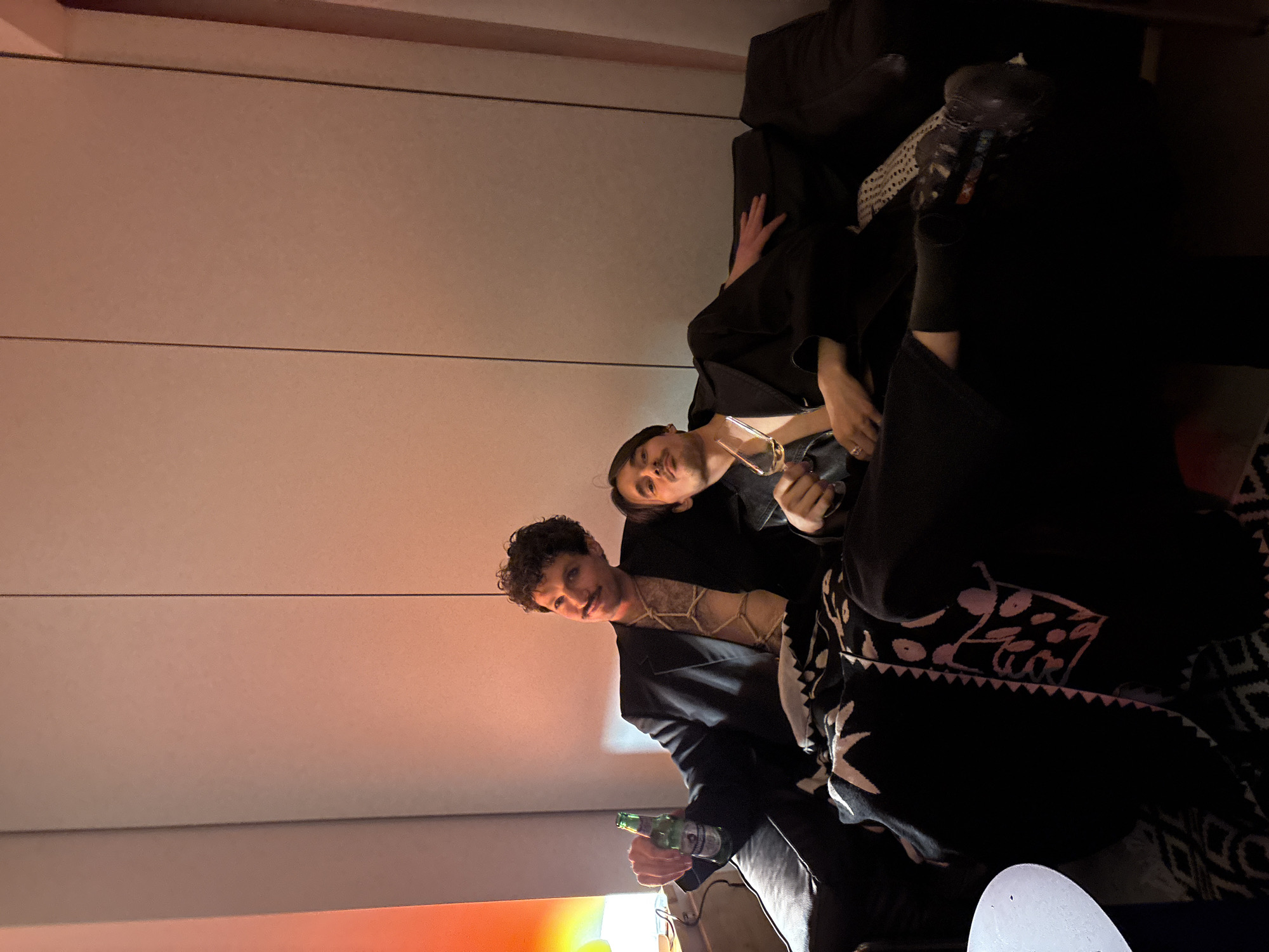 Two people are sitting on a black sofa indoors. One person holds a green beer bottle and wears a black jacket, a patterned top, and has curly dark hair. The other person is reclining, wearing black, and holding a glass. The background includes a light-colored wall with horizontal panels. The photo has a warm, orange-tinted lighting. The city of Moscow, Russia is not discernible from the provided image.