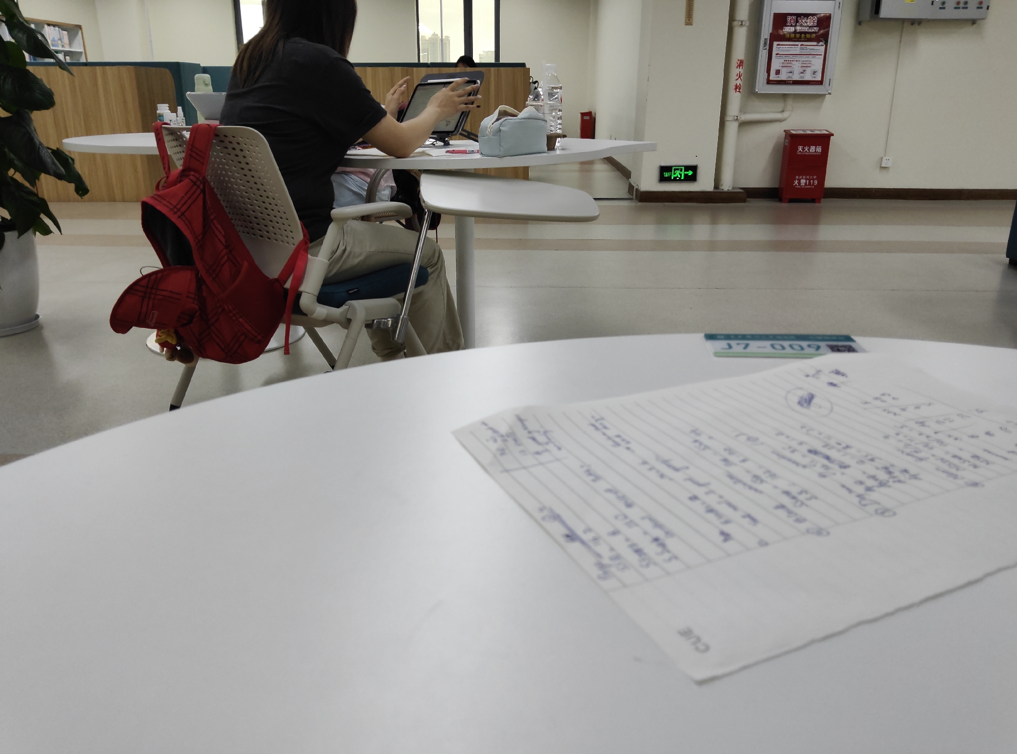This media file captures a scene within what appears to be a modern library or study space. The primary subject is a person, seen from behind and the side, seated at a white table and engaged with a tablet. They are wearing a dark top and light-colored pants. A red backpack with a plaid pattern is slung over the back of their chair. The setting is characterized by white, rounded tables and chairs, with a polished floor. In the background, there are glimpses of other study areas, including wooden panels and a large plant in a white pot. The lighting suggests it is daytime. Visible text includes signage in Chinese. One sign near a red fire extinguisher box reads "灭火器箱" (fire extinguisher box) and "火警119" (fire alarm 119). Another sign above it reads "消火栓" (fire hydrant). There is also a green "exit" sign with an arrow pointing to the right. A small card on the table in the foreground displays "J7-009". A piece of paper with handwritten notes in blue ink is also visible on the table in the foreground. The overall atmosphere seems to be one of quiet study or work. --- The user wants a description of the provided image. I should focus on the elements present in the image and the text that is visible. **Subjects:** * **People:** One person, seen from behind and side, using a tablet. * **Objects:** Red plaid backpack, white tables, chairs, tablet, water bottle, pencil case, fire extinguisher box, fire hydrant sign, exit sign, a card with text "J7-009", and a paper with handwritten notes. * **Scene/Setting:** Interior, likely a library, study area, or communal workspace. Modern design with white furniture, polished floors, and some wooden elements. * **Activity:** The person is studying or working on a tablet. **Notable Details:** * **Location Cues:** Chinese text on signage related to fire safety (fire hydrant, fire extinguisher, fire alarm number 119). Green exit sign. * **Time of Day:** Indoor lighting suggests daytime. * **Emotions:** Neutral, suggesting focus or concentration. **Visible Text:** * "→" * "消火栓" (fire hydrant) * "消火栓" (fire hydrant) * "FIRE HAIDRANT" * "消防安全知识" (fire safety knowledge) * "灭火器箱" (fire extinguisher box) * "火警119" (fire alarm 119) * "J7-009" * "CUE" I will structure the description logically, starting with the main subject and scene, then elaborating on details and visible text. I will aim for conciseness.This media file captures an indoor scene,