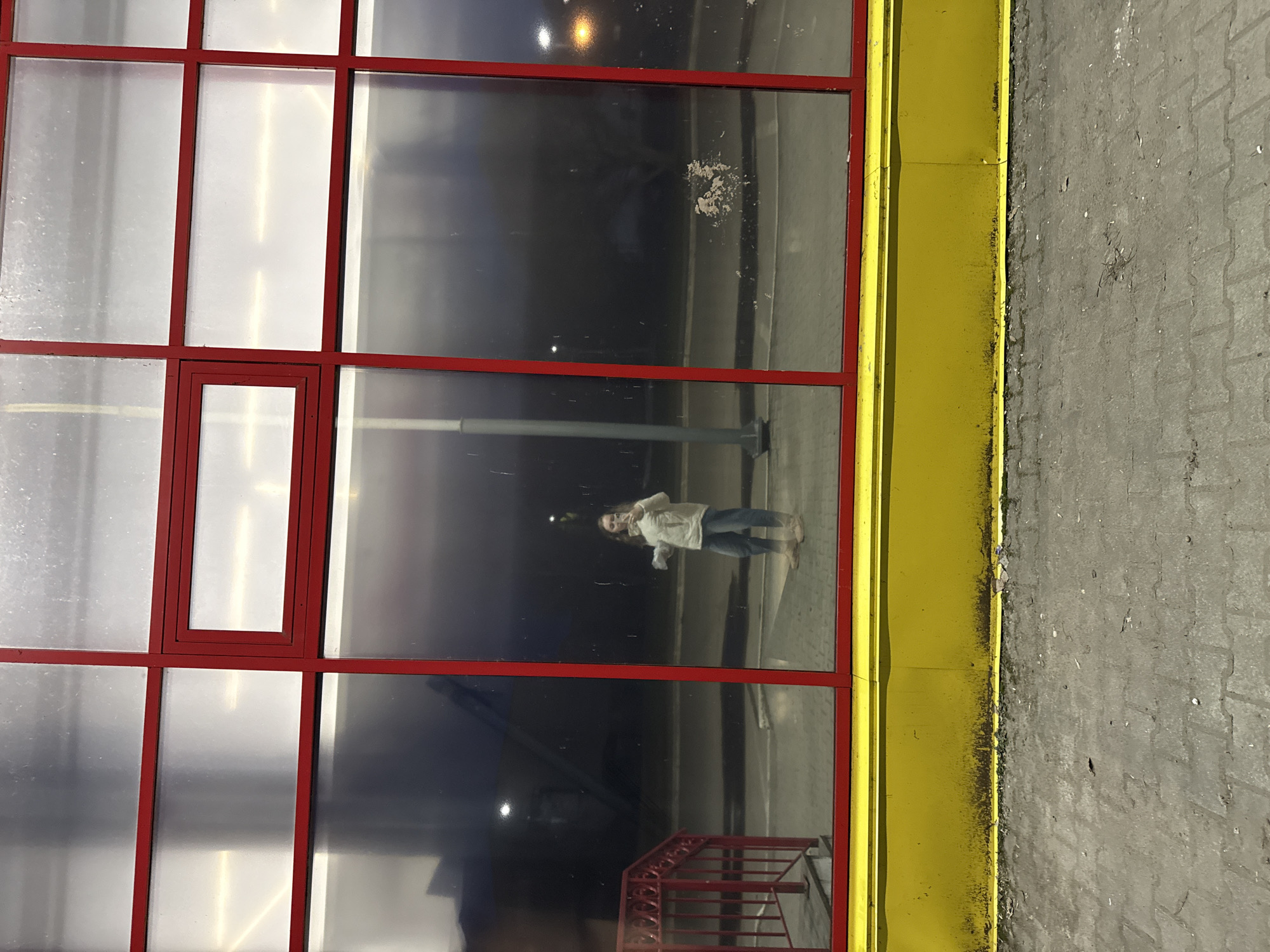 A medium shot depicts a reflection of a person in a window. The window, framed with red borders and set in a yellow structure, is a grid pattern with multiple panes. The reflection of the person, dressed in light-colored clothing and jeans, is seen in the center-right of the window's reflection. The scene is in Unknown, Unknown.