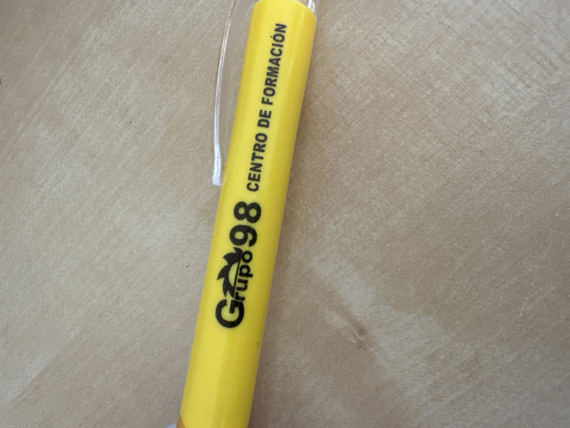 The image is a close-up, top-down shot of a yellow pen resting on a beige, textured surface, likely a table or countertop. The pen has black text and a logo printed on it. The text reads "Grupo 98" followed by a stylized symbol and then "CENTRO DE FORMACIÓN". There are no people or any indication of the city of Murcia, Spain, present in the image. The lighting is natural and casts subtle shadows.