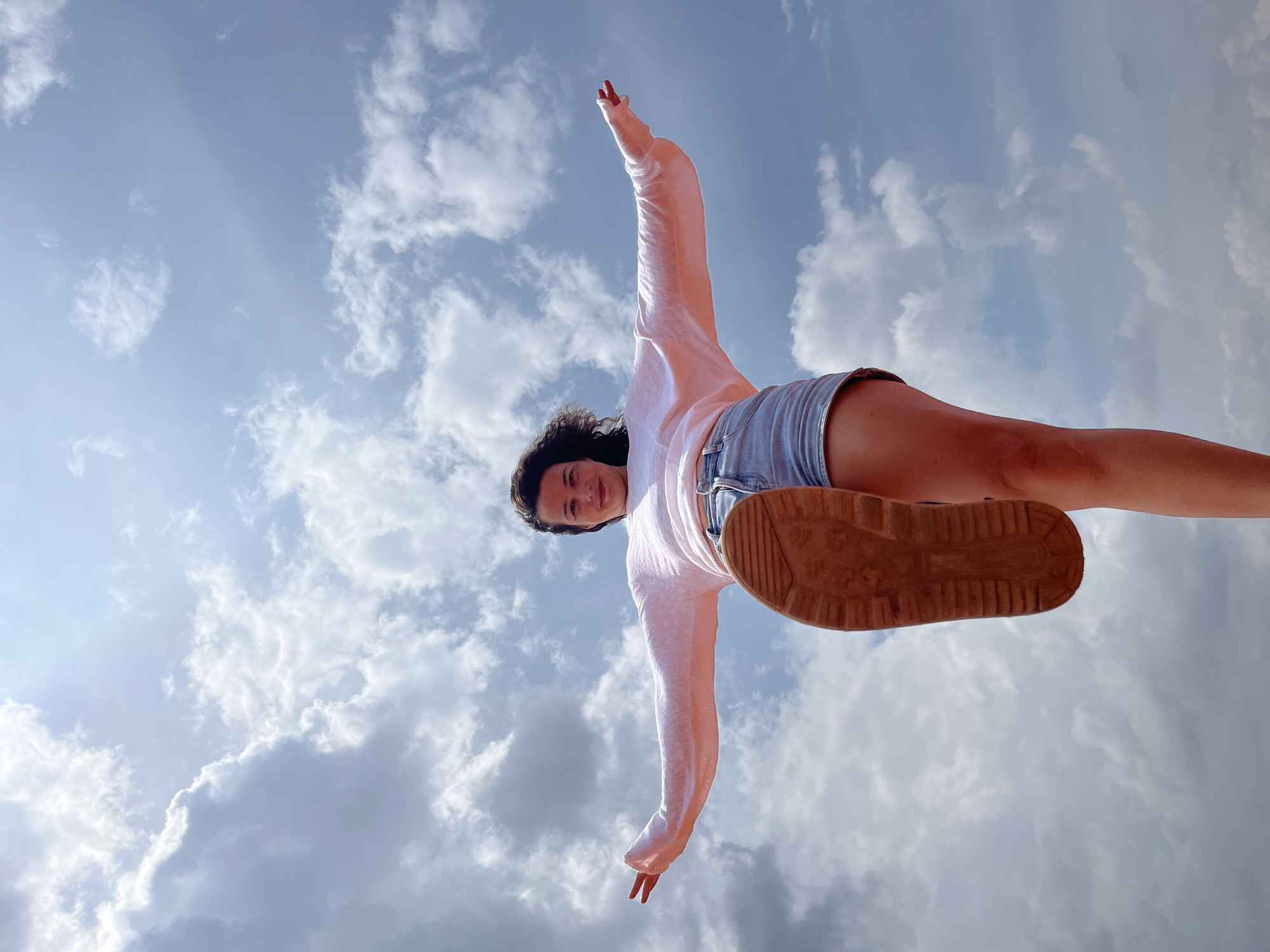 A low-angle, medium shot captures a young woman standing against a bright, cloudy sky, her right leg extended forward and her left arm outstretched to the side, palm open. She is wearing a light pink long-sleeved shirt and denim shorts. The sole of her right shoe is prominently visible, angled towards the viewer. The background consists of a dynamic sky filled with fluffy white clouds and patches of blue. The scene appears to be outdoors. This image was taken in Mui Ne, Vietnam.