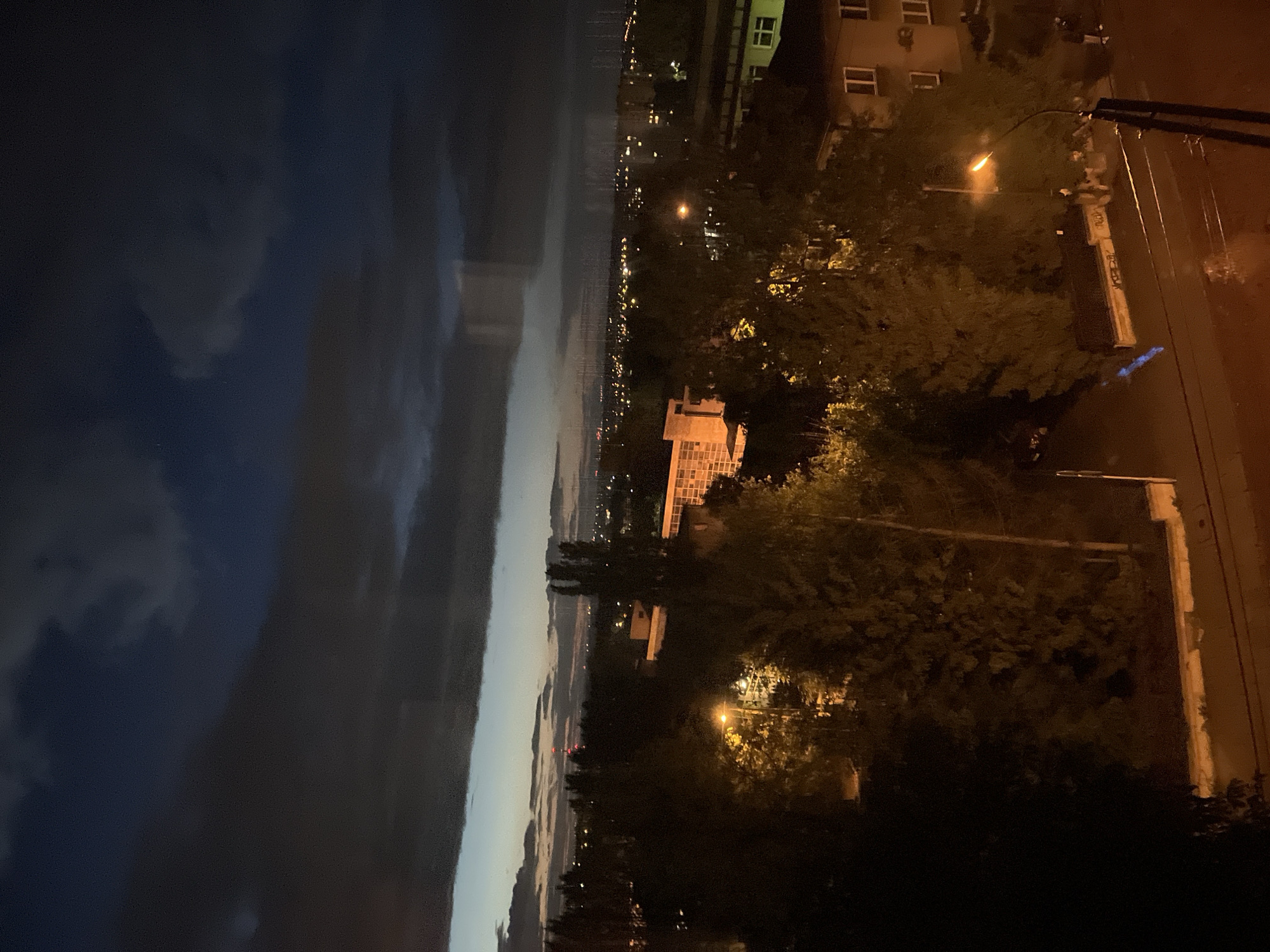 An elevated night-time wide shot captures a cityscape under a dark, cloudy sky. The foreground is dominated by trees and a lamppost illuminating a street. A building with a light-colored facade and windows is visible to the left of the frame, partially obscured by trees. In the mid-ground, several more buildings with illuminated windows are scattered throughout the scene, interspersed with more trees. The background shows a distant city skyline with numerous lights, suggesting a populated area. The sky is a deep blue and black, with heavy clouds casting a shadow over the city. The overall impression is one of a quiet, dimly lit urban environment as dusk settles.