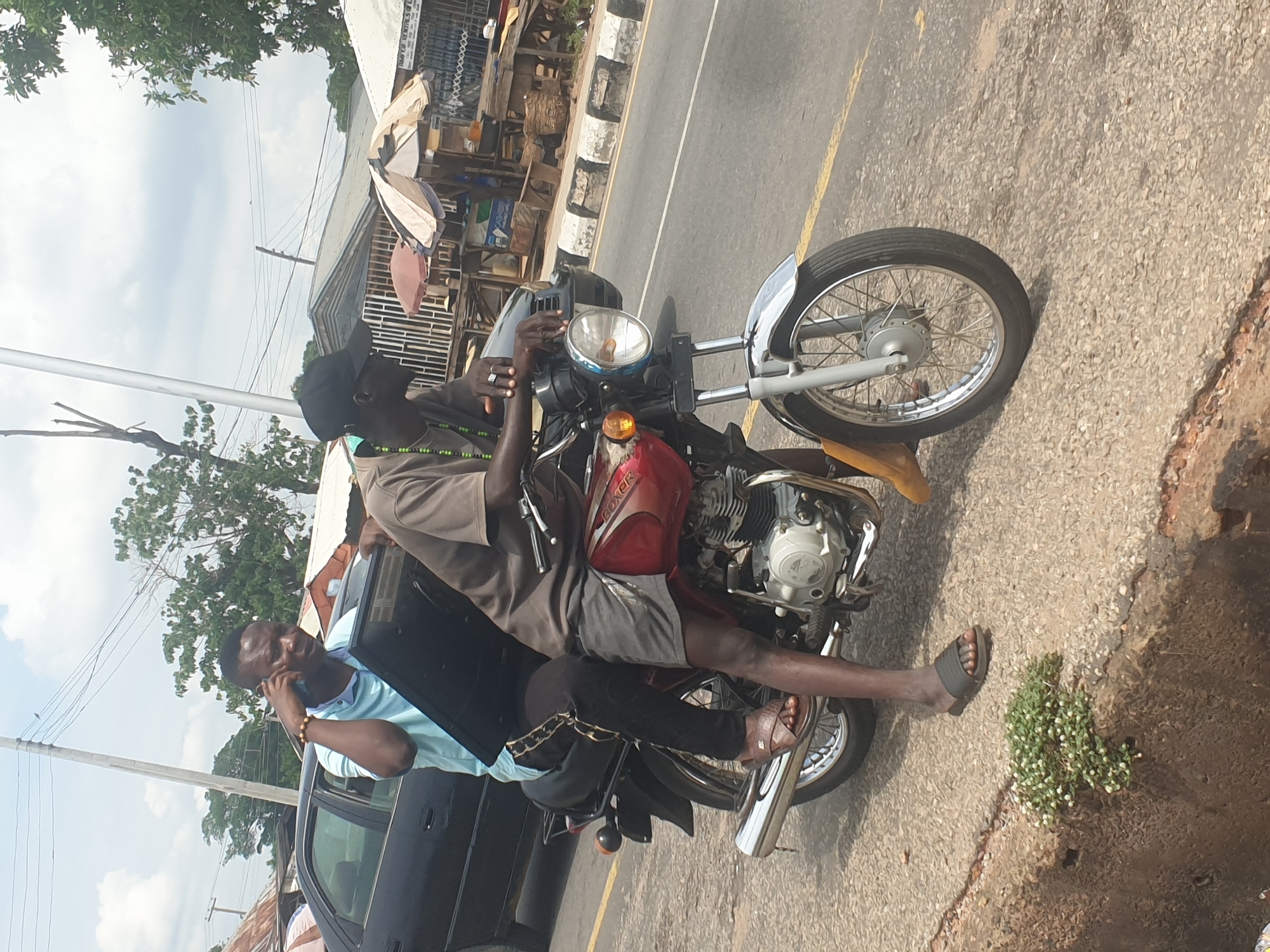 Two men are visible, one sitting on a red motorcycle while looking at his phone, and another one leaning on a dark vehicle, talking on his phone. The scene appears to be on a street in an urban setting. The surroundings include buildings, trees, and street infrastructure.